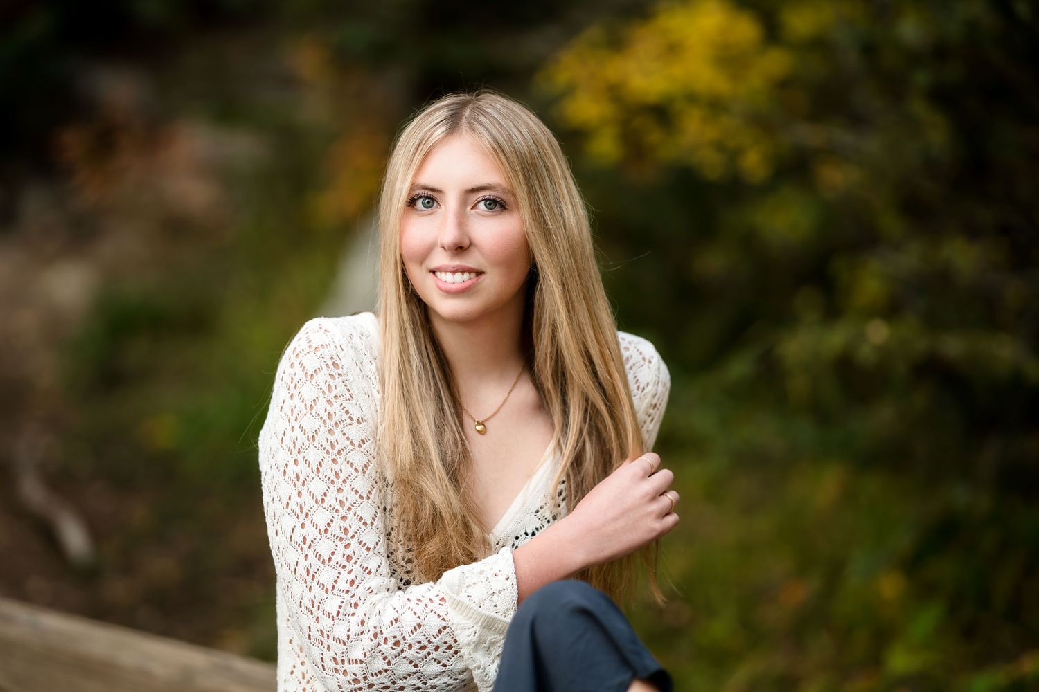 Young blonde woman smiling outdoors in white lace top, seated against a lush green and yellow foliage background.