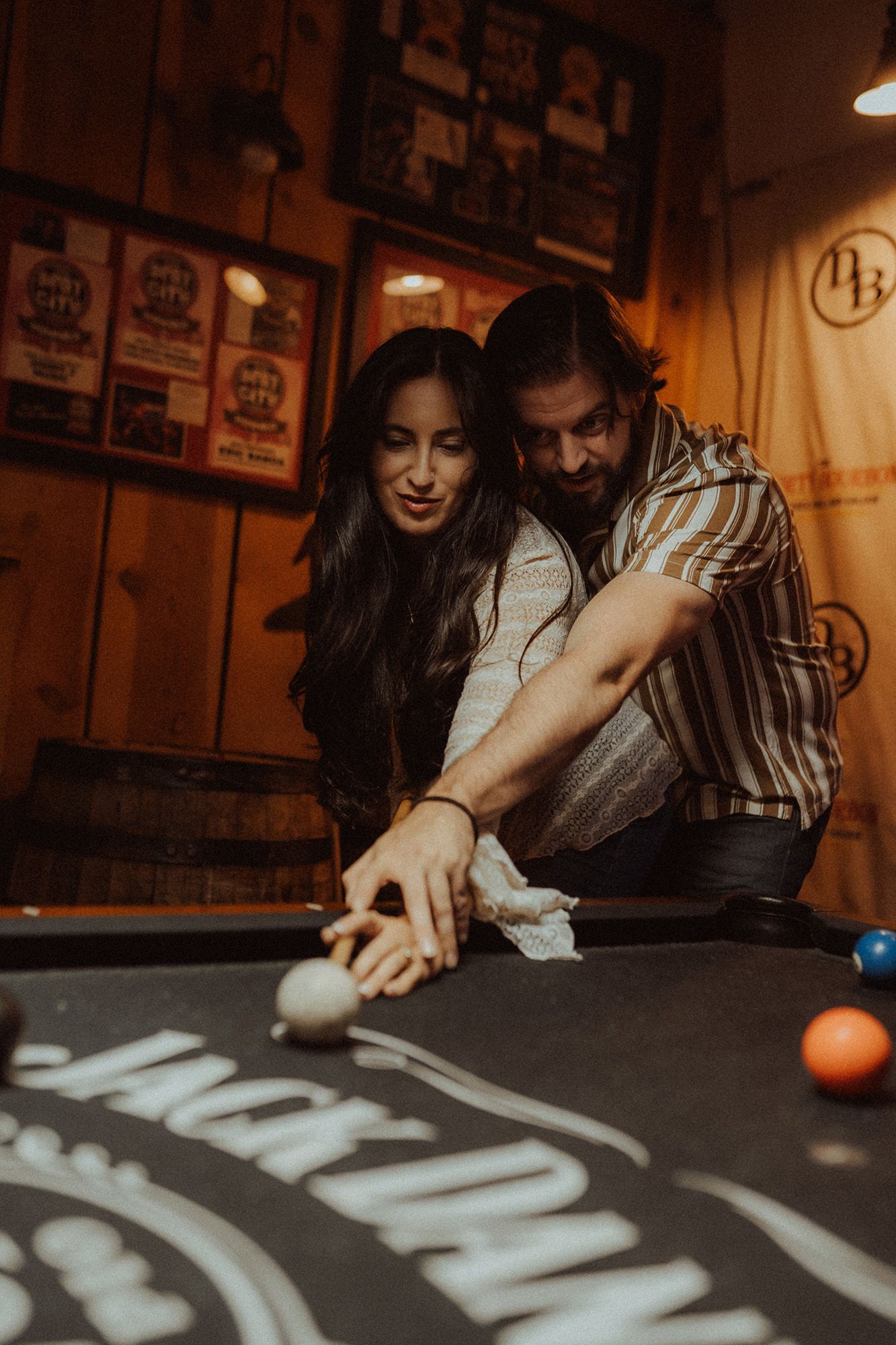 Couple playing pool together at a bar, man guiding woman's shot on a Jack Daniels billiard table.