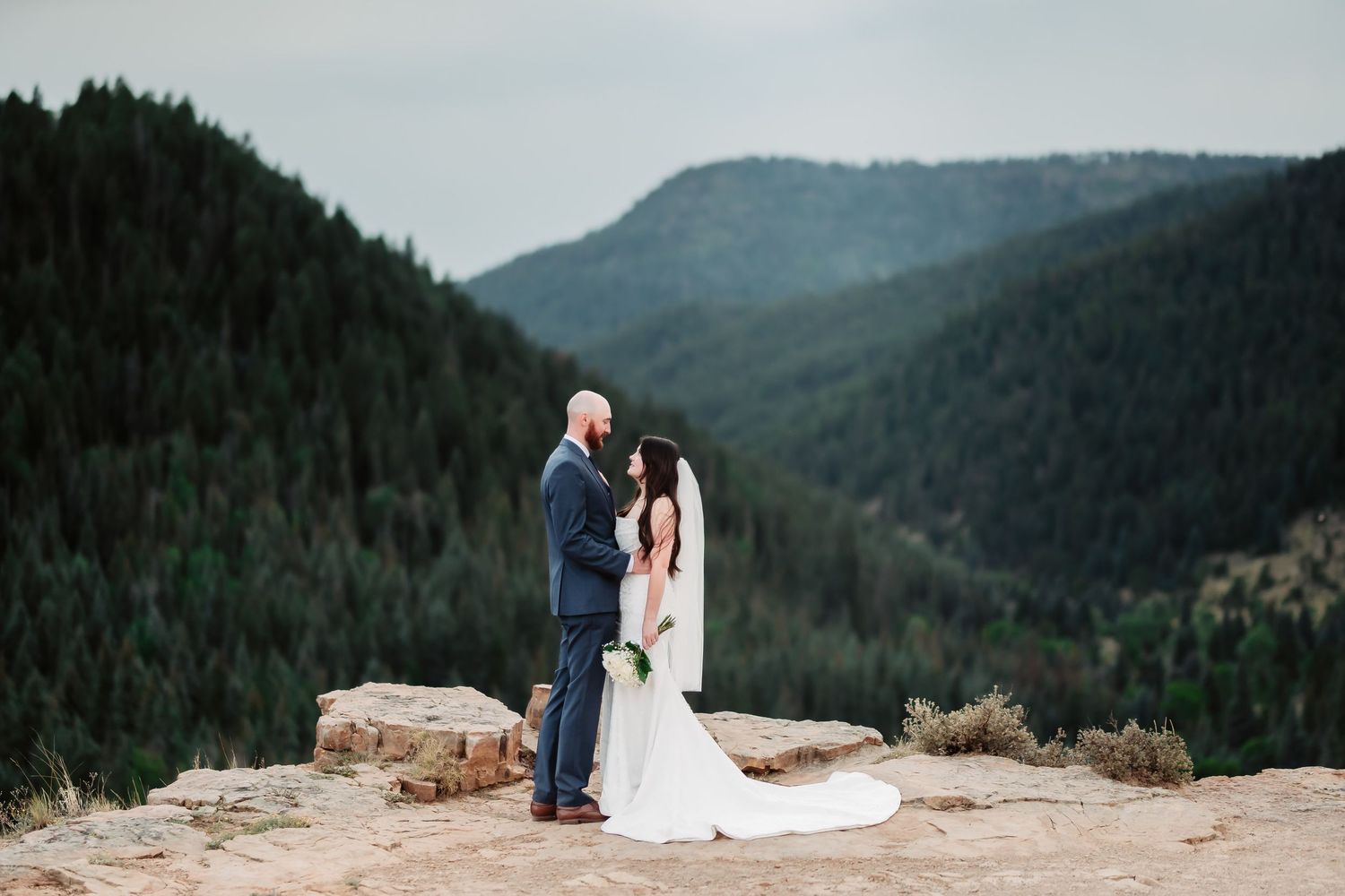 A couple in wedding attire stands on a mountain cliff overlooking a scenic forested valley landscape at sunset.