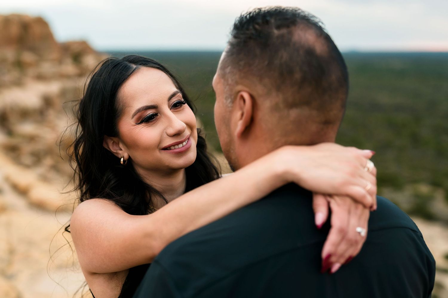 Smiling woman with dark wavy hair embracing her partner at a scenic desert overlook with sandstone cliffs.