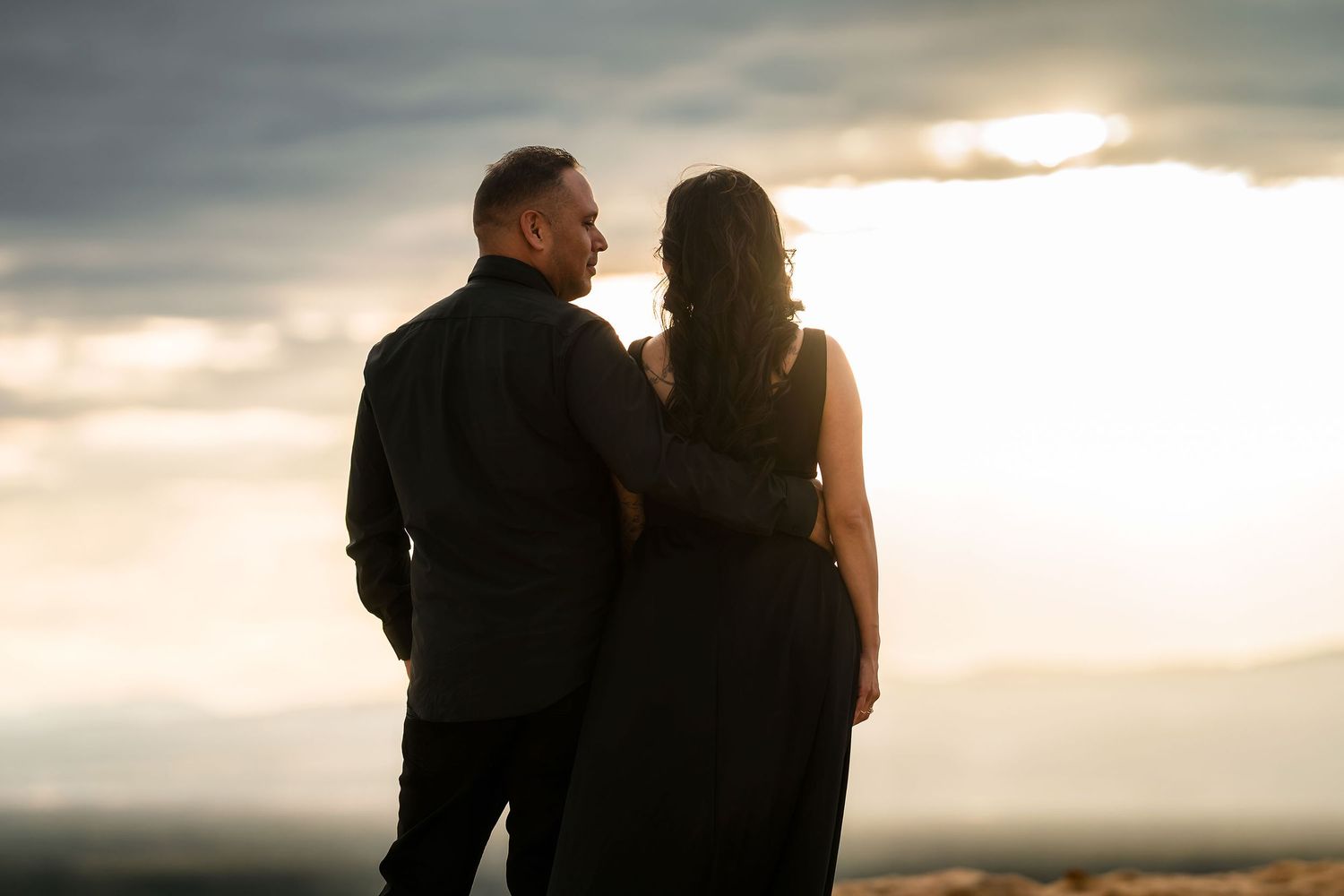 Romantic couple in black silhouetted against glowing sunset sky sharing an intimate moment outdoors.