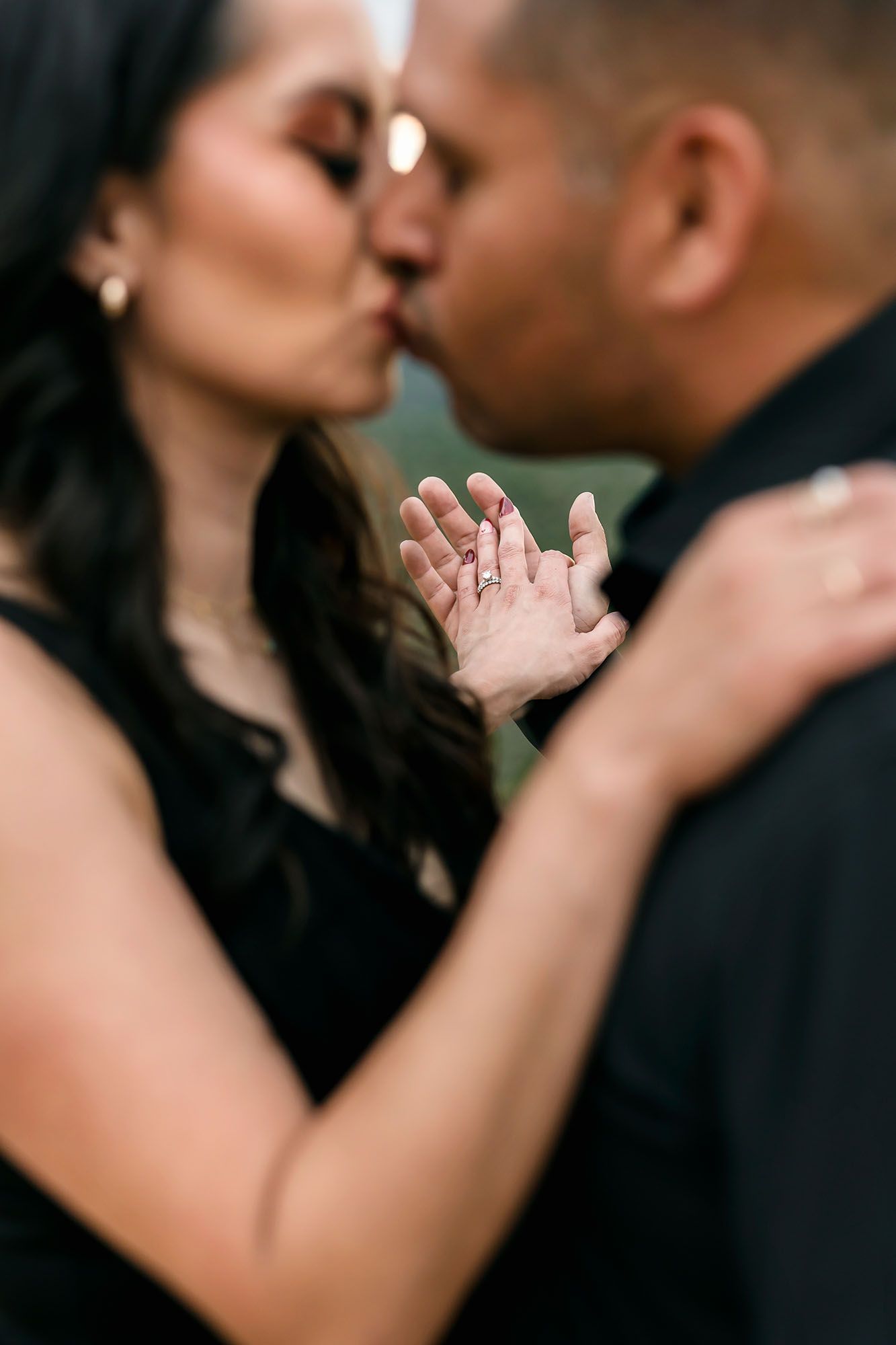 Engaged couple kissing while woman displays diamond ring on open palm in soft focus portrait.