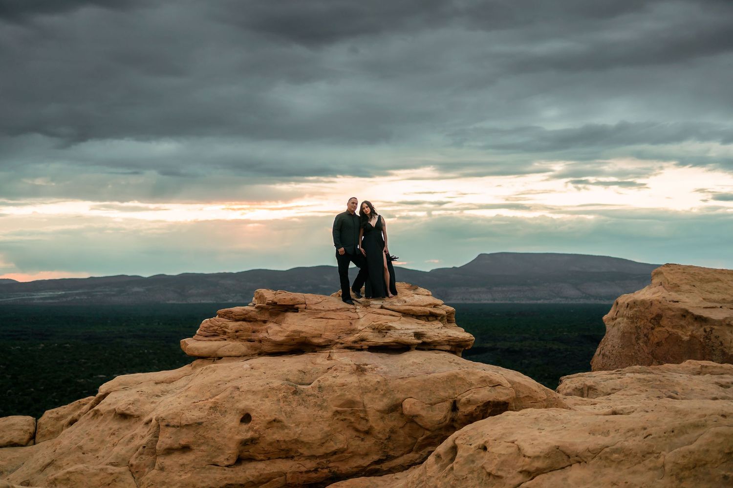 Couple standing together on rocky overlook with stunning panoramic mountain views under moody storm clouds.