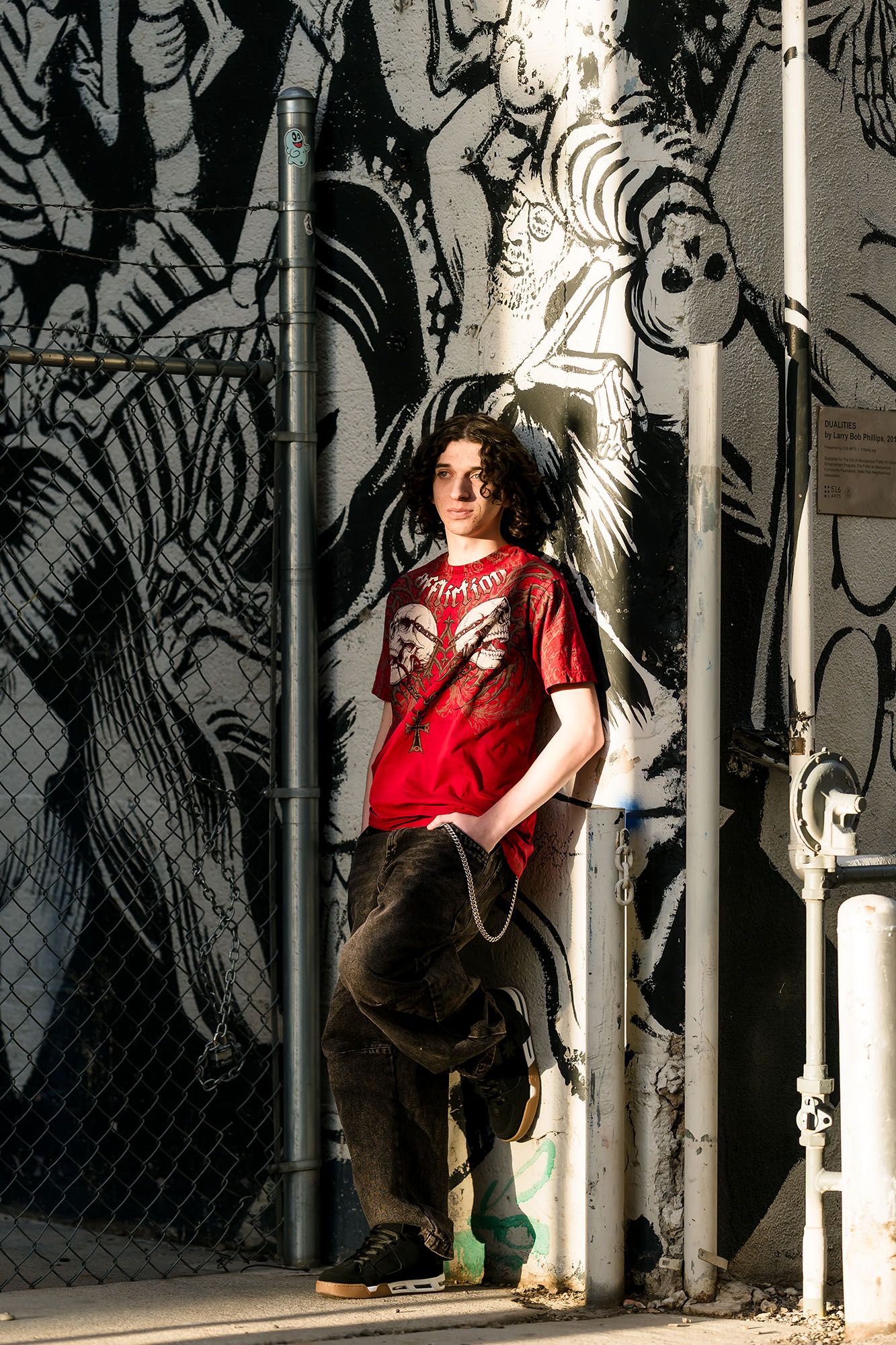 Young person in red graphic tee poses against black and white street art mural near chain-link fence.