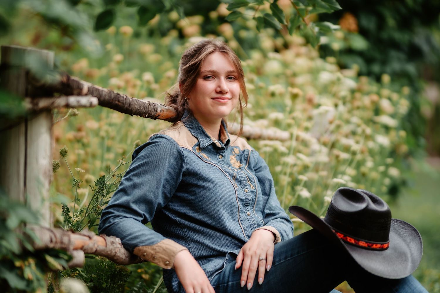 Person in denim shirt poses outdoors in a garden setting with natural greenery and foliage in the background.