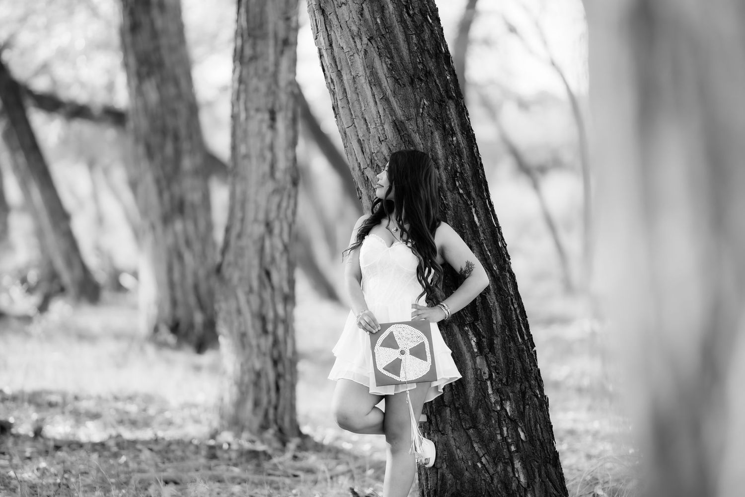 Black and white portrait of graduate posing near tree trunk during celebration photoshoot.
