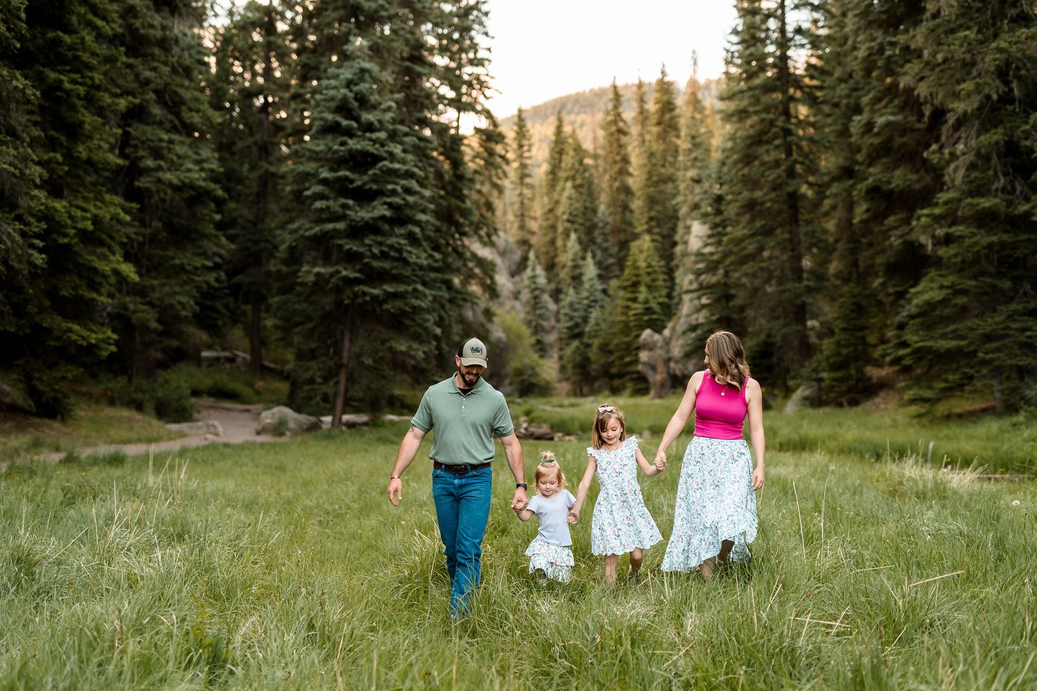 Family of four holding hands walking through a lush green mountain meadow surrounded by tall pine trees.
