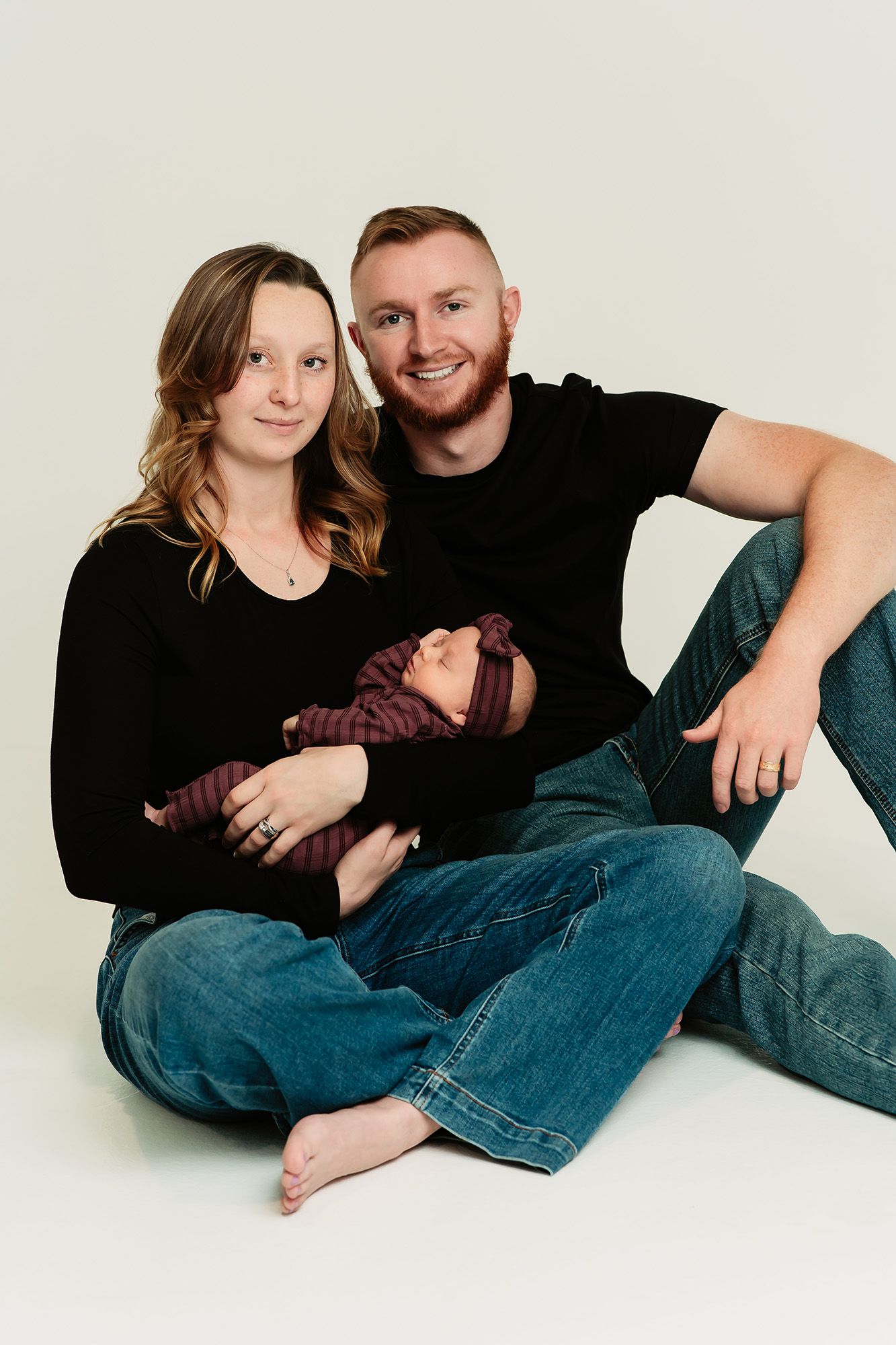 A young couple in black tops and jeans sit together on a white backdrop, lovingly cradling their newborn baby.