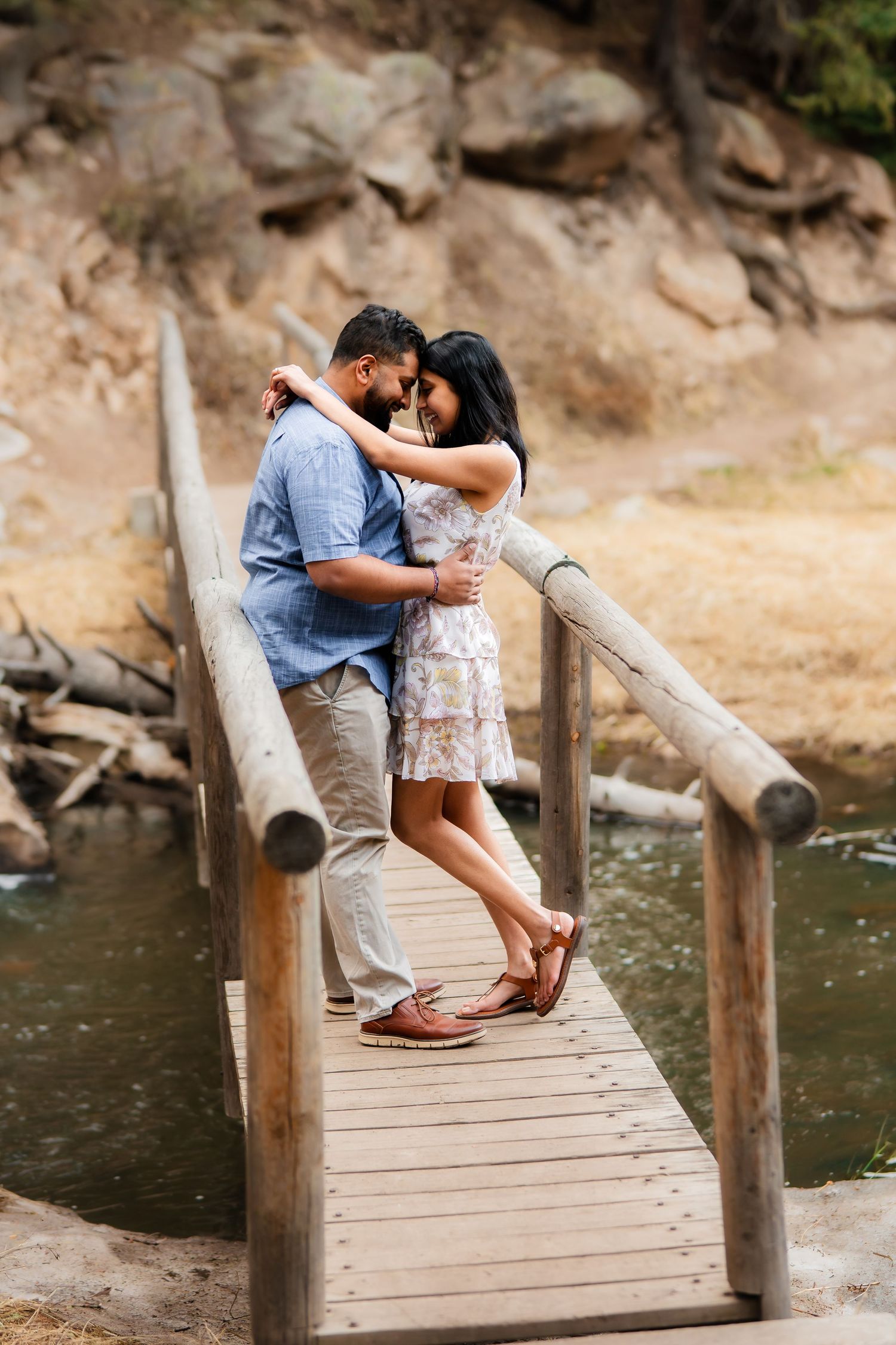 A romantic couple embraces on a wooden footbridge over water in a scenic outdoor setting.