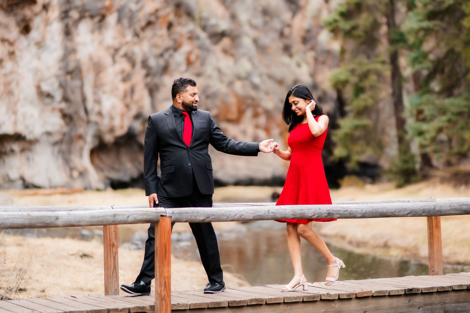 A playful scene on a rustic bridge with a couple in red and black formal wear.