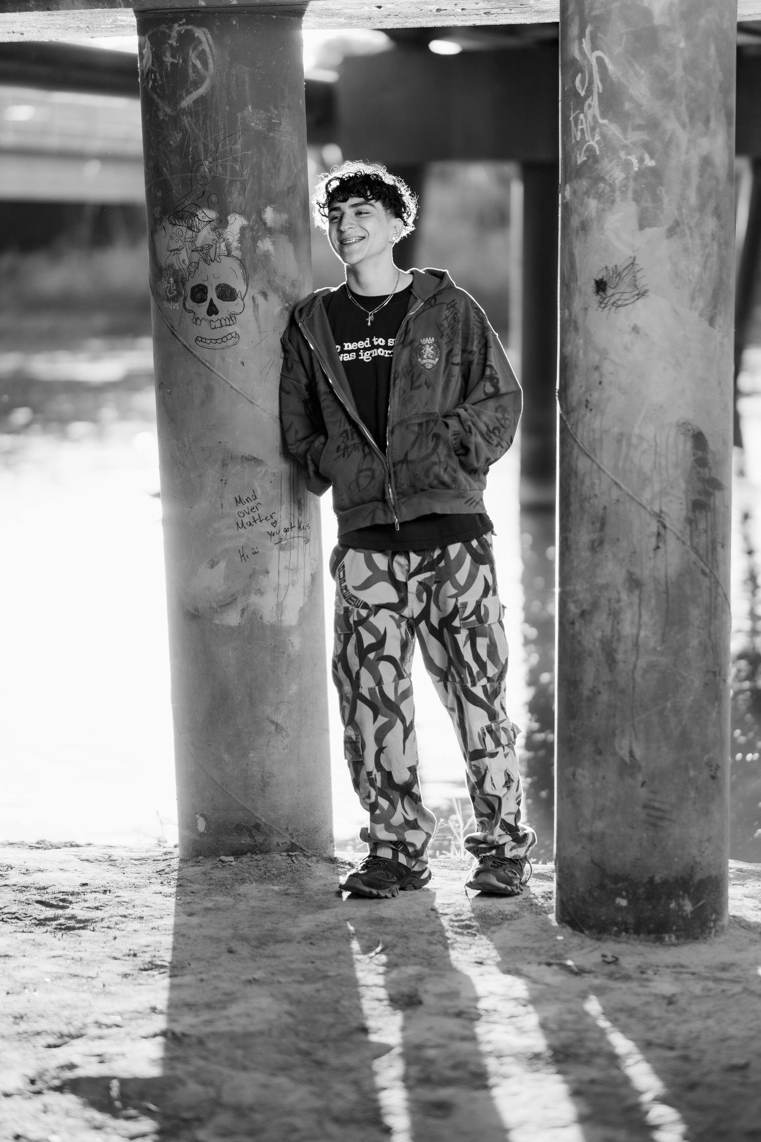 Black and white photo of person in casual streetwear standing between concrete pillars with graffiti.