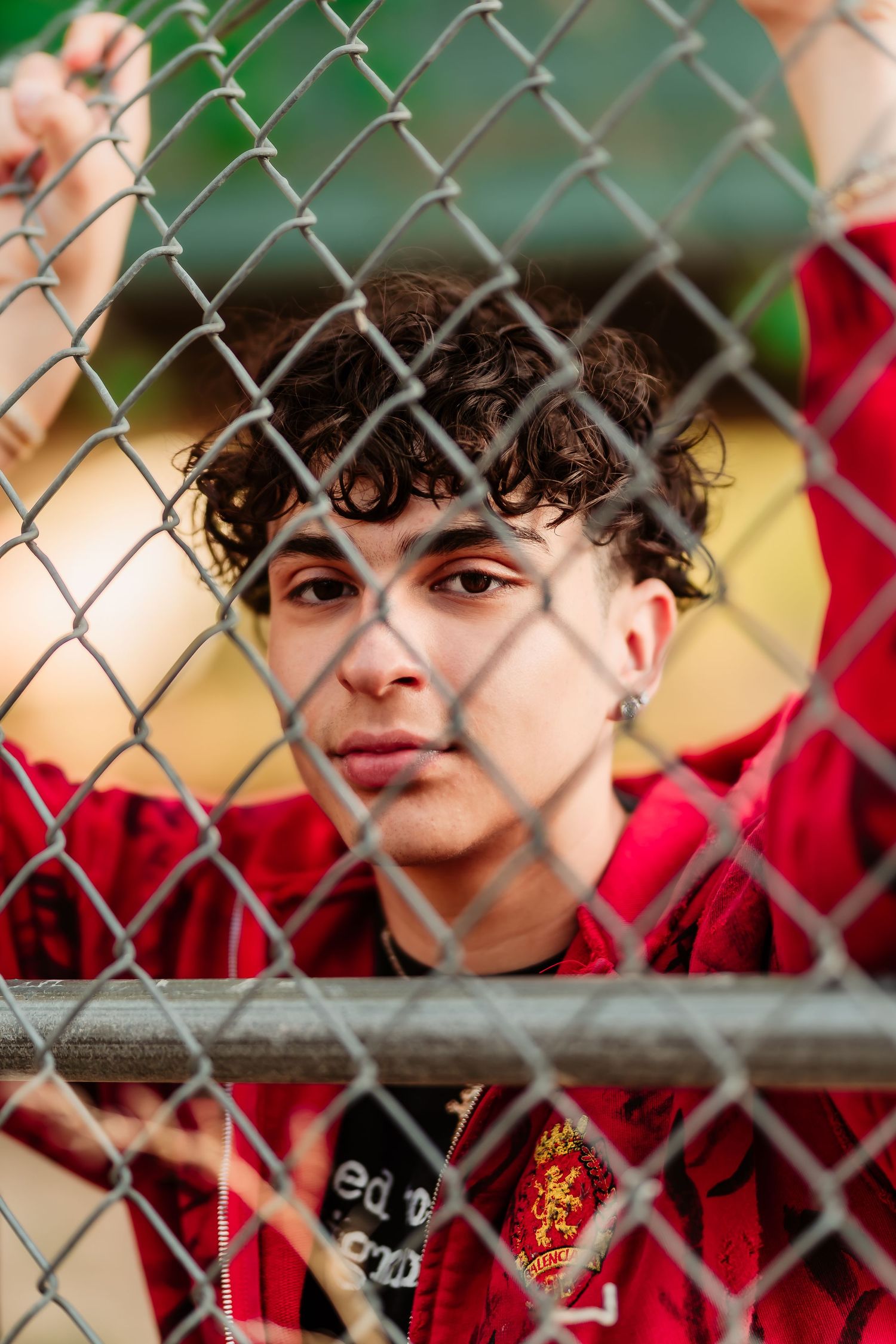 A close-up portrait through a chain-link fence shows someone wearing a red top with curly hair.