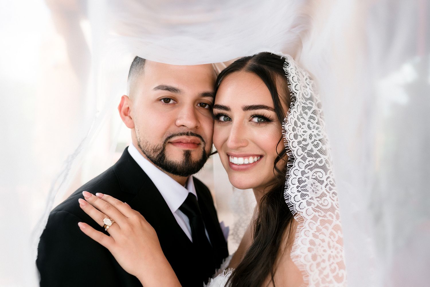 A smiling bride and groom pose closely together under a flowing white veil on their wedding day.