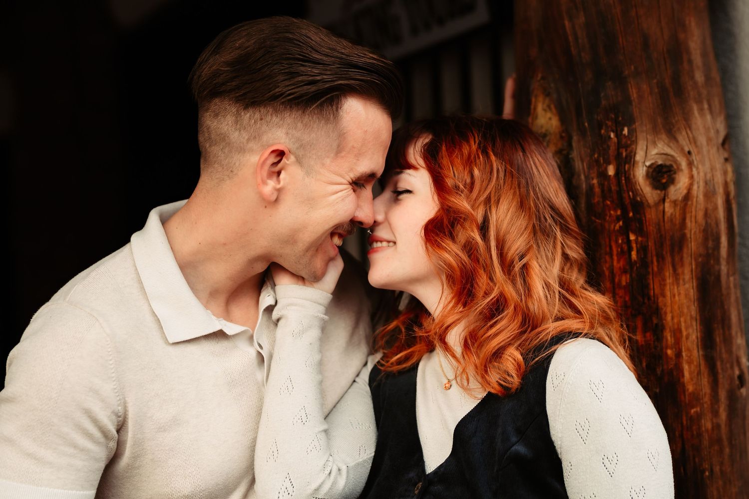 A romantic couple shares intimate moments against a dark wooden backdrop in warm lighting.
