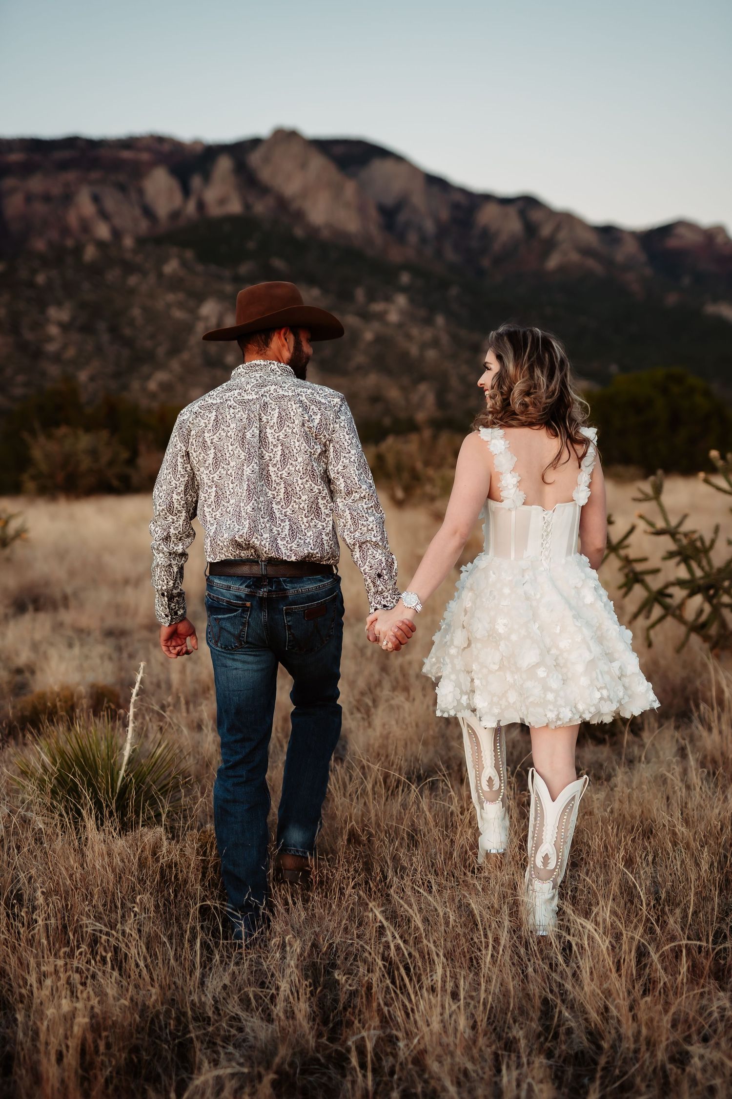 A couple walks hand in hand through a desert landscape with mountains in the background during golden hour.