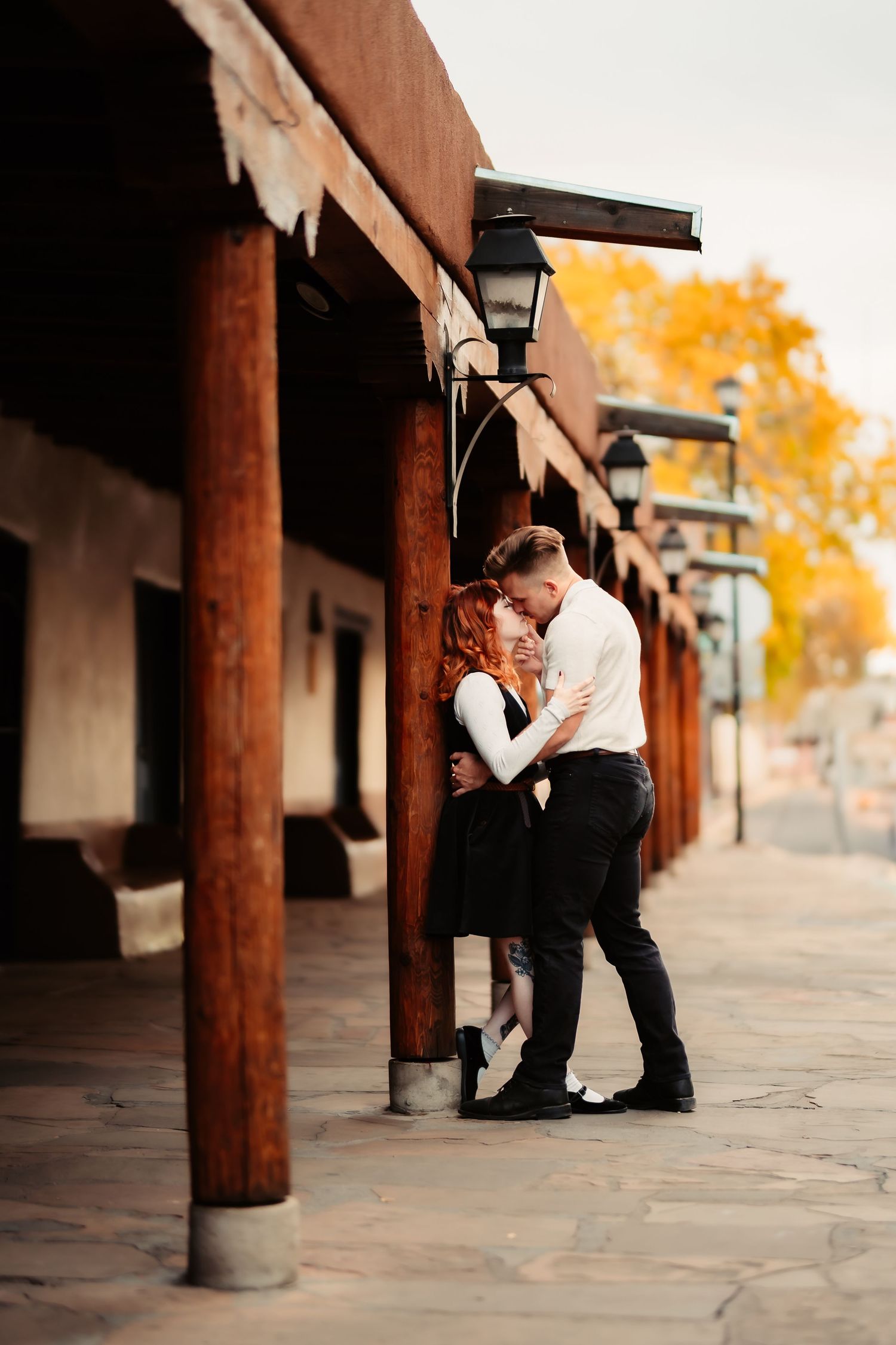 A romantic couple embraces under rustic wooden posts and lanterns along a covered walkway with autumn colors in the background.