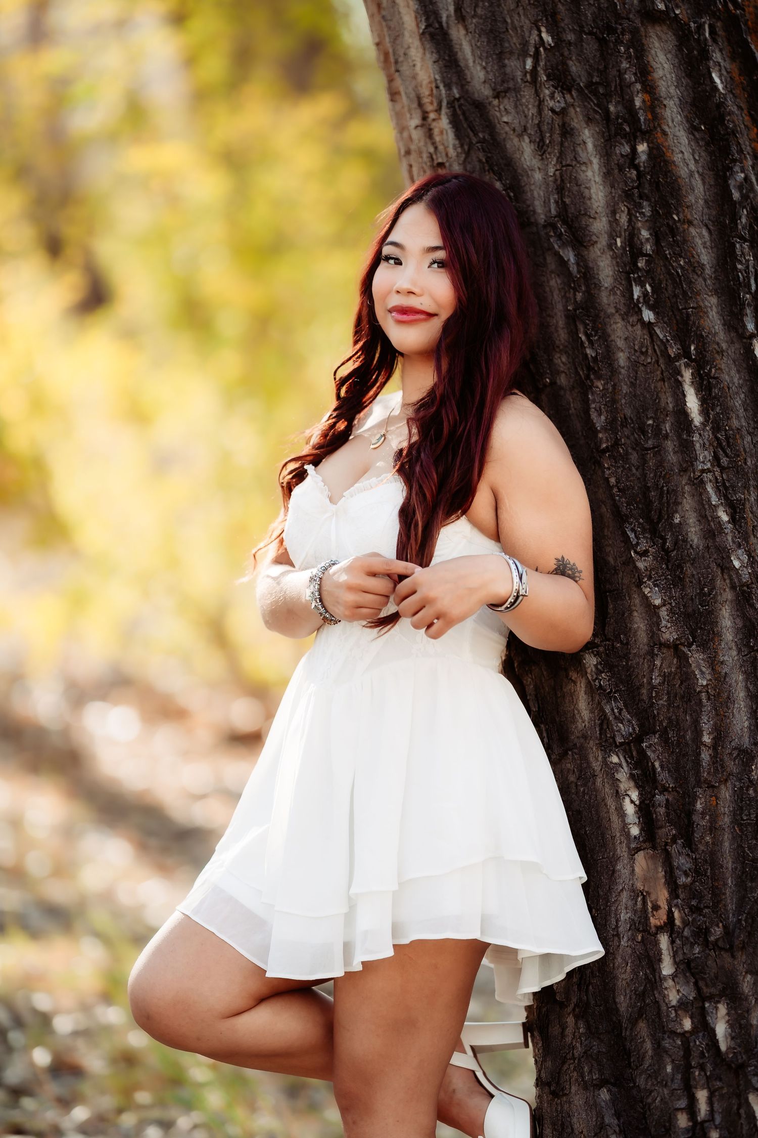 Outdoor maternity photoshoot in white dress with autumn foliage and tree backdrop.