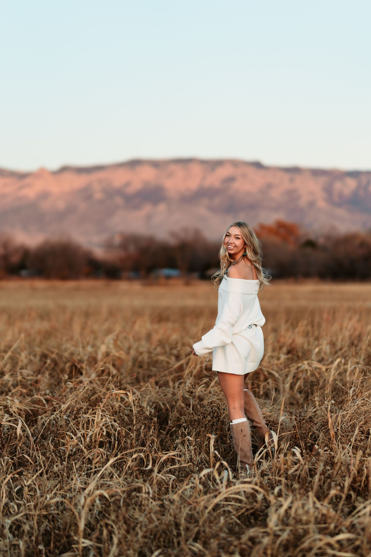 A white off-shoulder dress styled with boots against a mountain landscape in warm autumn light.