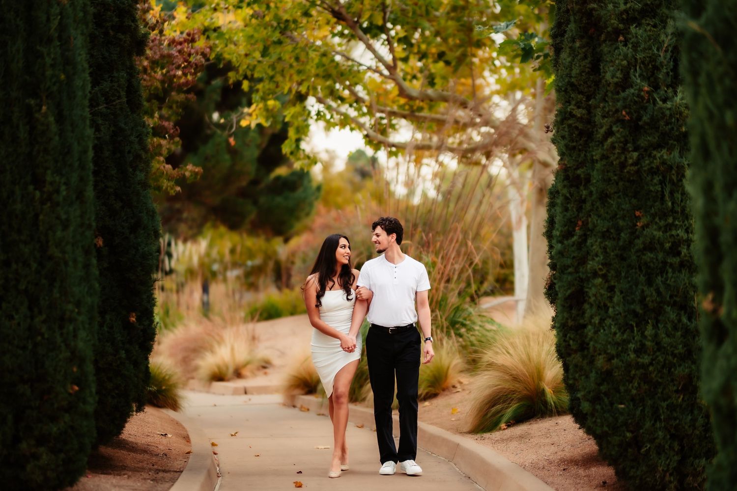 A couple walks along a tree-lined pathway during a romantic autumn engagement photo session.