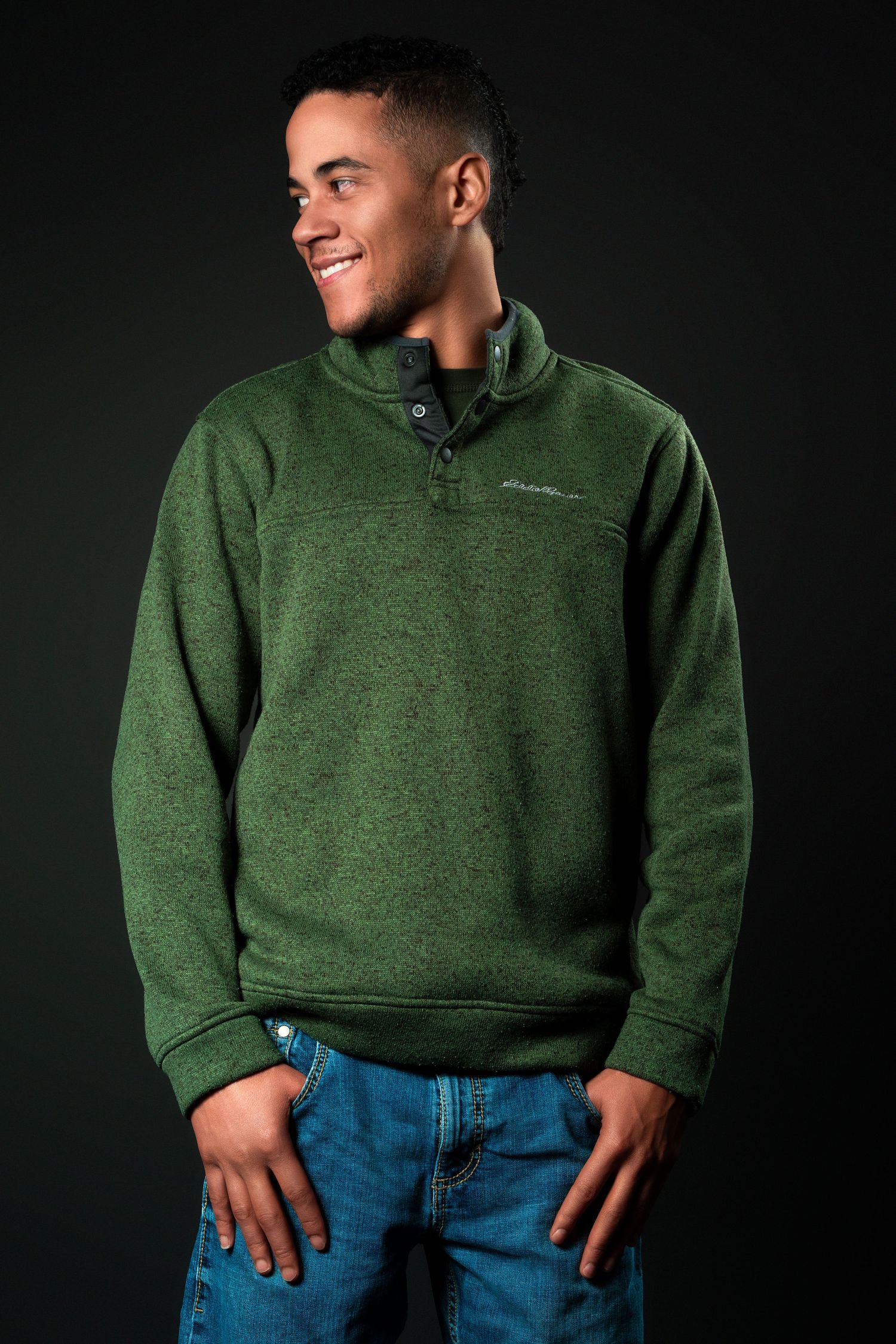 A person wearing a green fleece pullover sweater and blue jeans poses against a dark background.