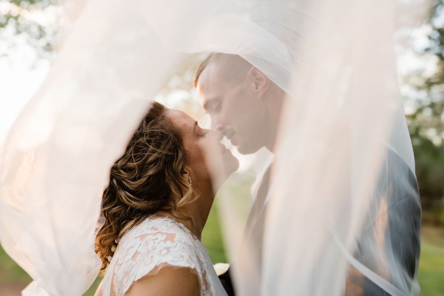 A romantic wedding couple shares a tender kiss under a flowing white veil in an outdoor garden setting.