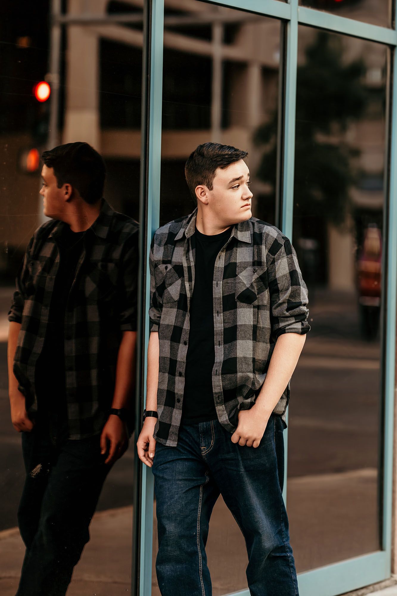 Young man in plaid flannel shirt leaning against glass building reflecting city street at dusk.
