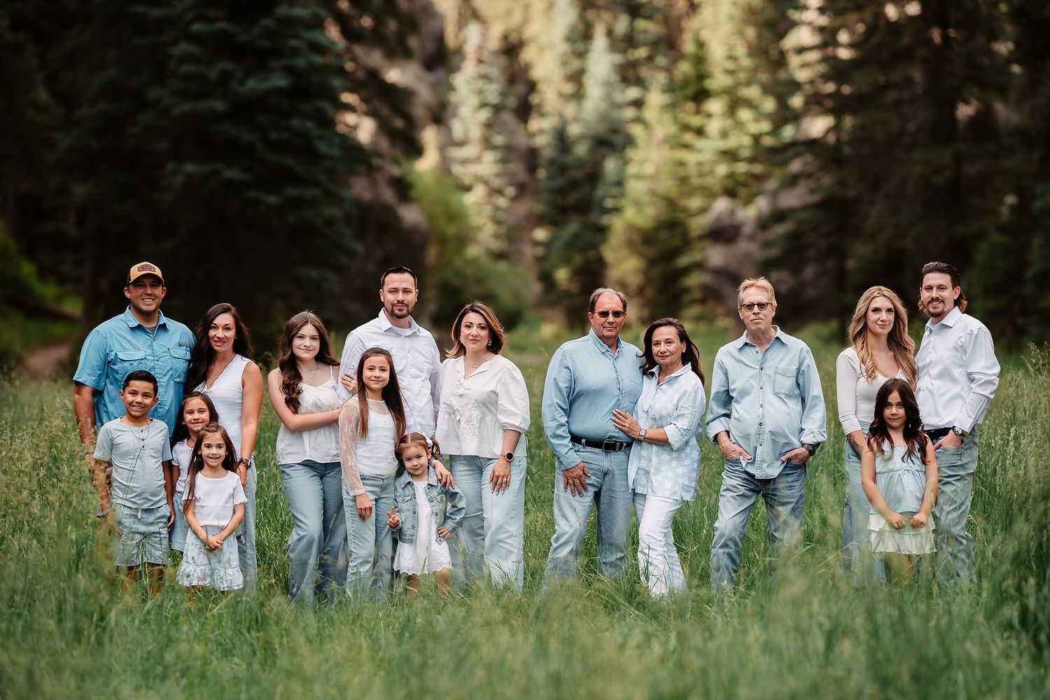 Large family group portrait outdoors in a lush green meadow surrounded by tall pine trees, wearing coordinated casual outfits.
