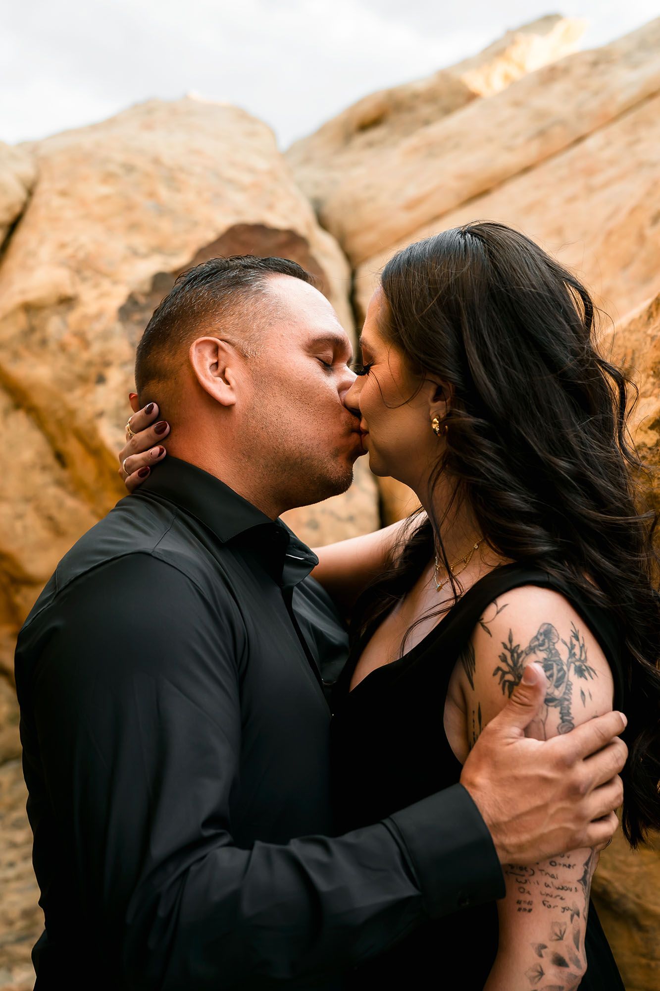 Couple sharing a romantic kiss against golden sandstone rocks, woman with tattoos in black dress.