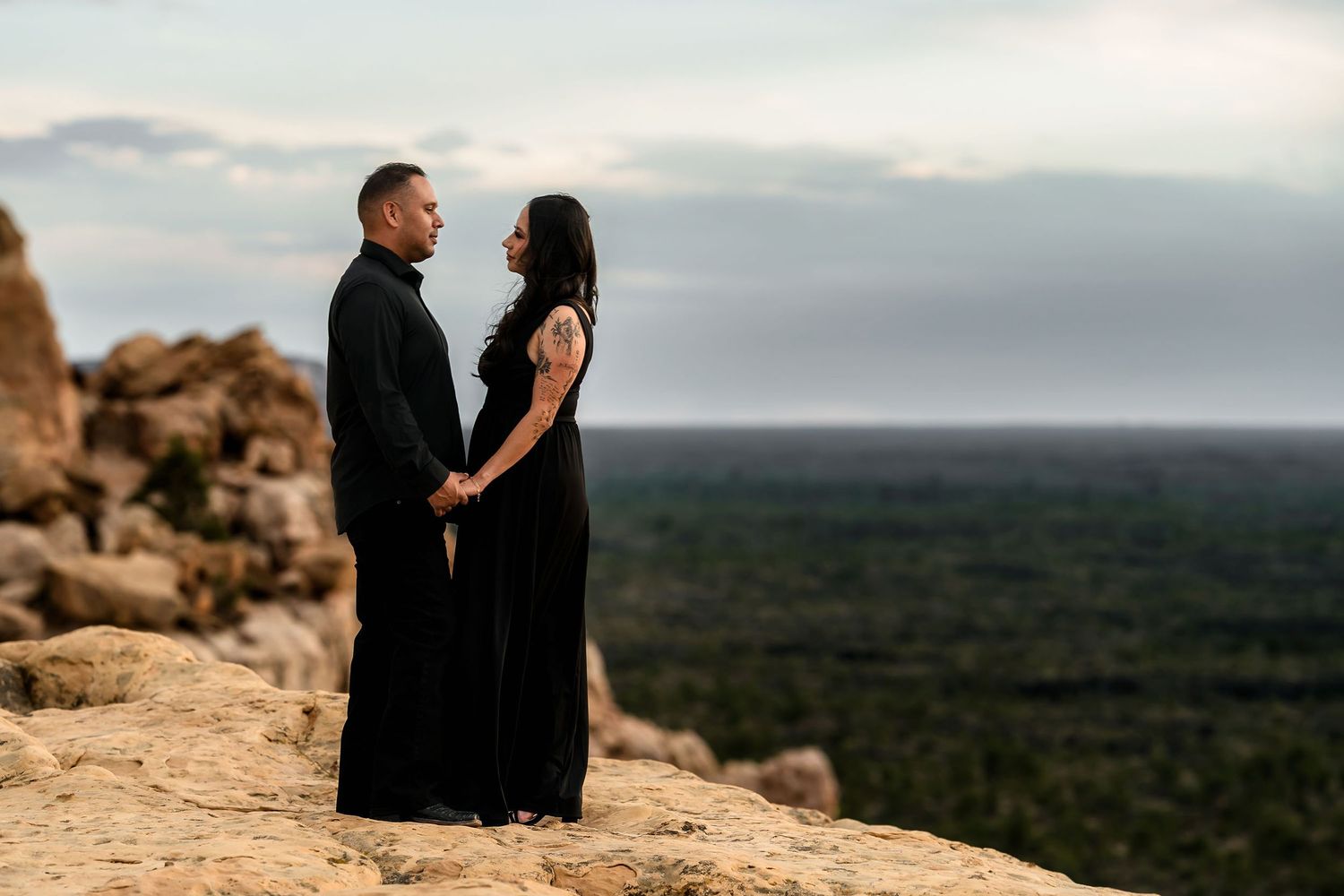 Couple in matching black outfits holding hands on rocky cliff with vast landscape at sunset.