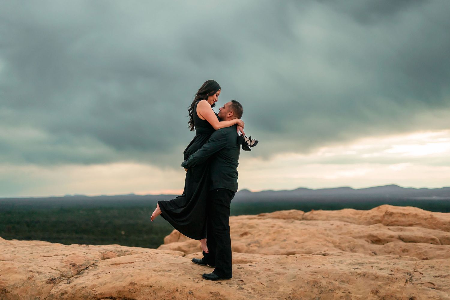 Man lifting woman in black dress on rocky cliff edge with moody overcast sky backdrop.