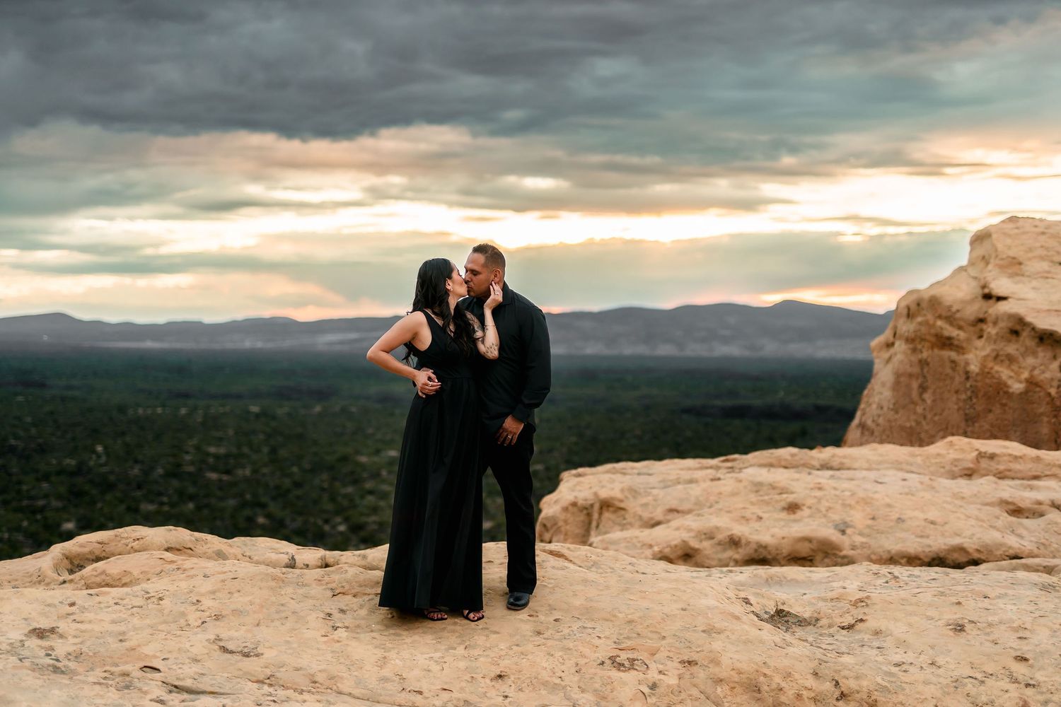 Couple in matching black outfits kissing on rocky cliff at sunset with mountain views.