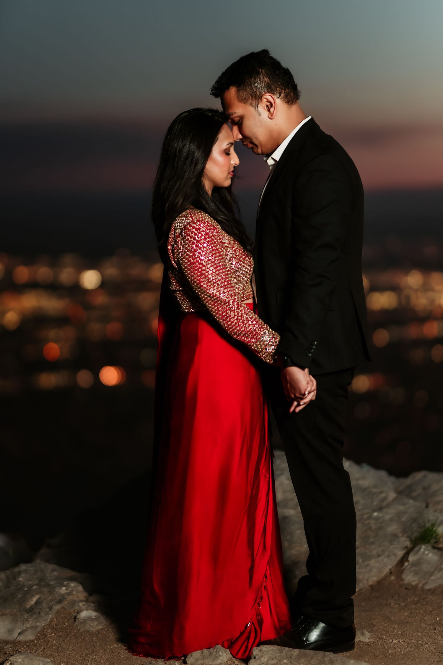 Romantic couple in red dress and black suit pose for sunset engagement photos against a city skyline backdrop.