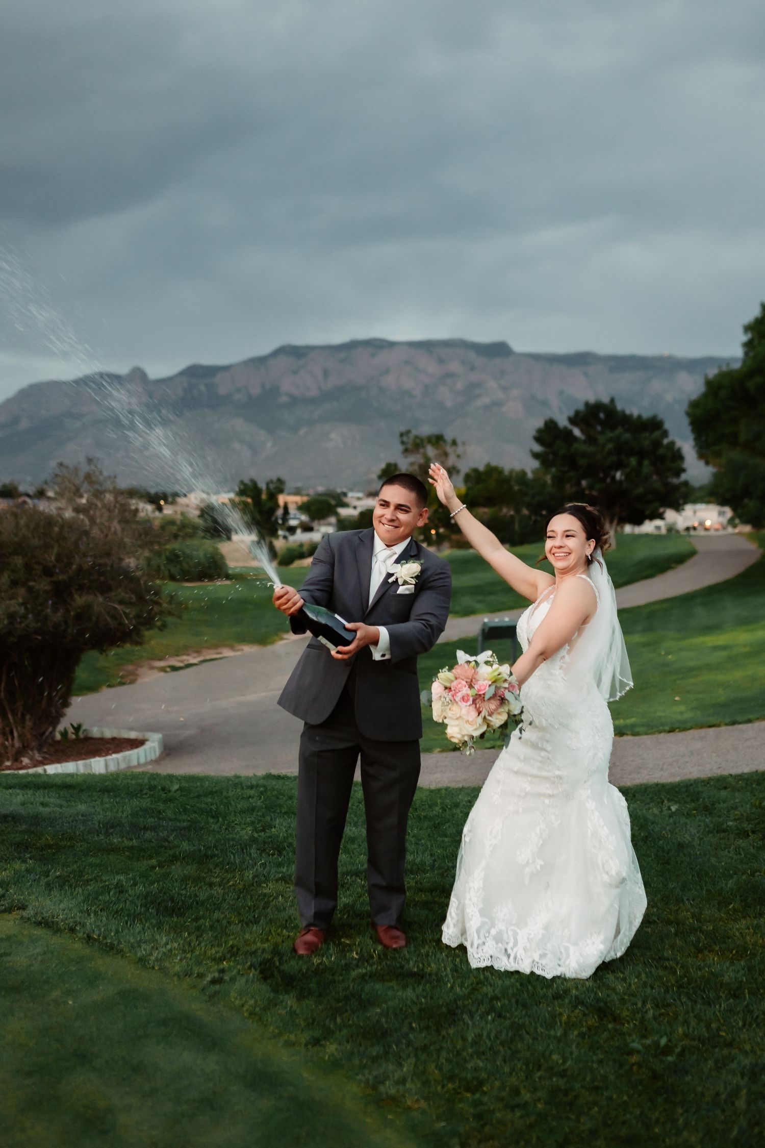 Playful wedding couple poses together on grassy hill with dramatic cloudy mountain backdrop.