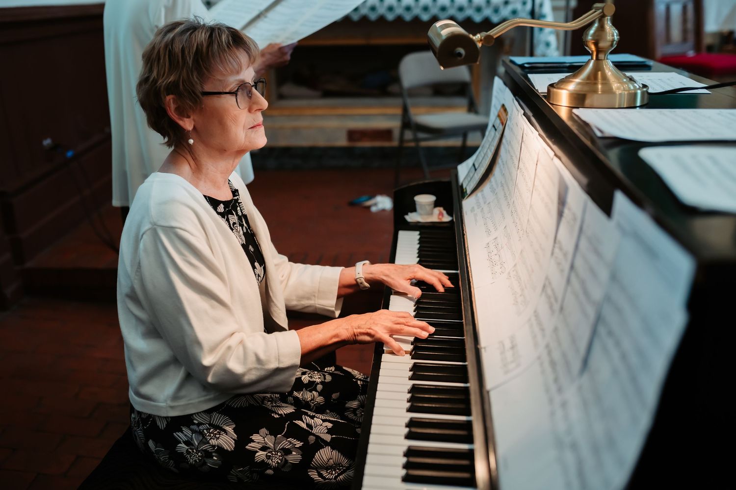 Person wearing glasses and white top plays piano keyboard while seated at mirrored glass window in home studio setting.