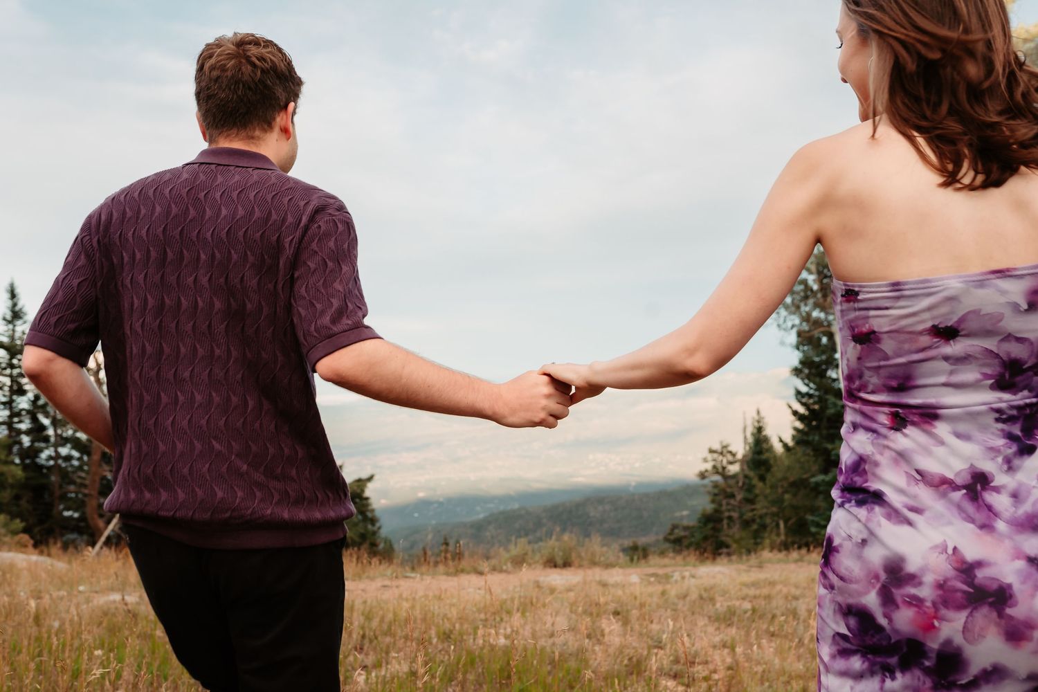 Holding hands while walking through a mountain meadow with evergreen trees in background.