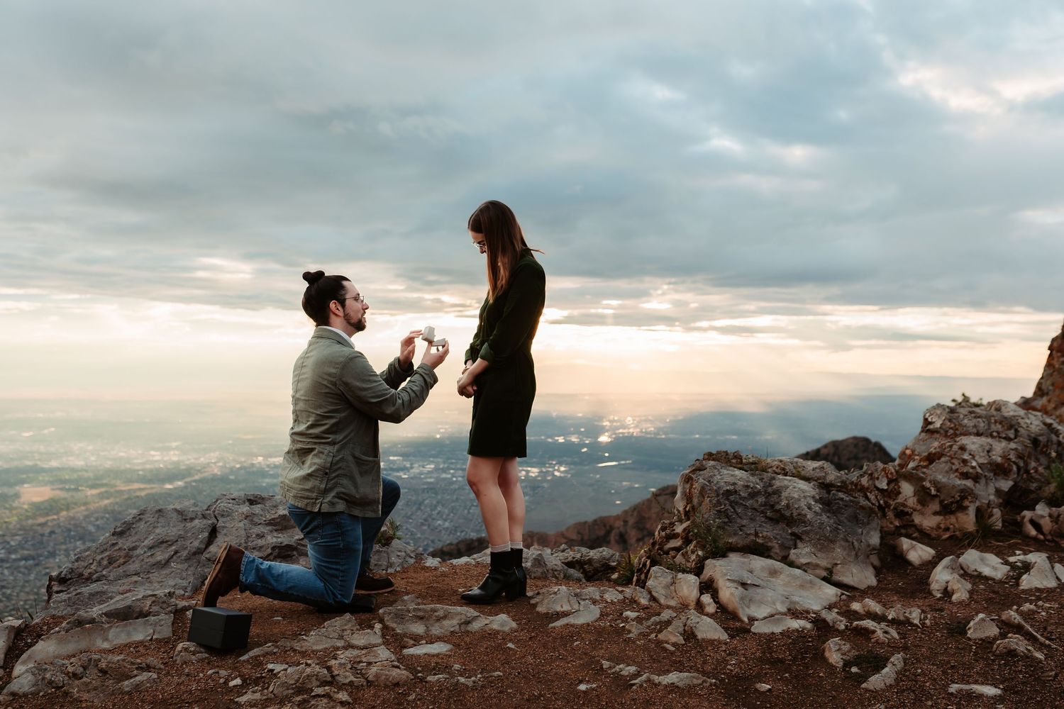 Two people kneel during a romantic proposal moment atop a mountain summit against a cloudy sky during sunset.
