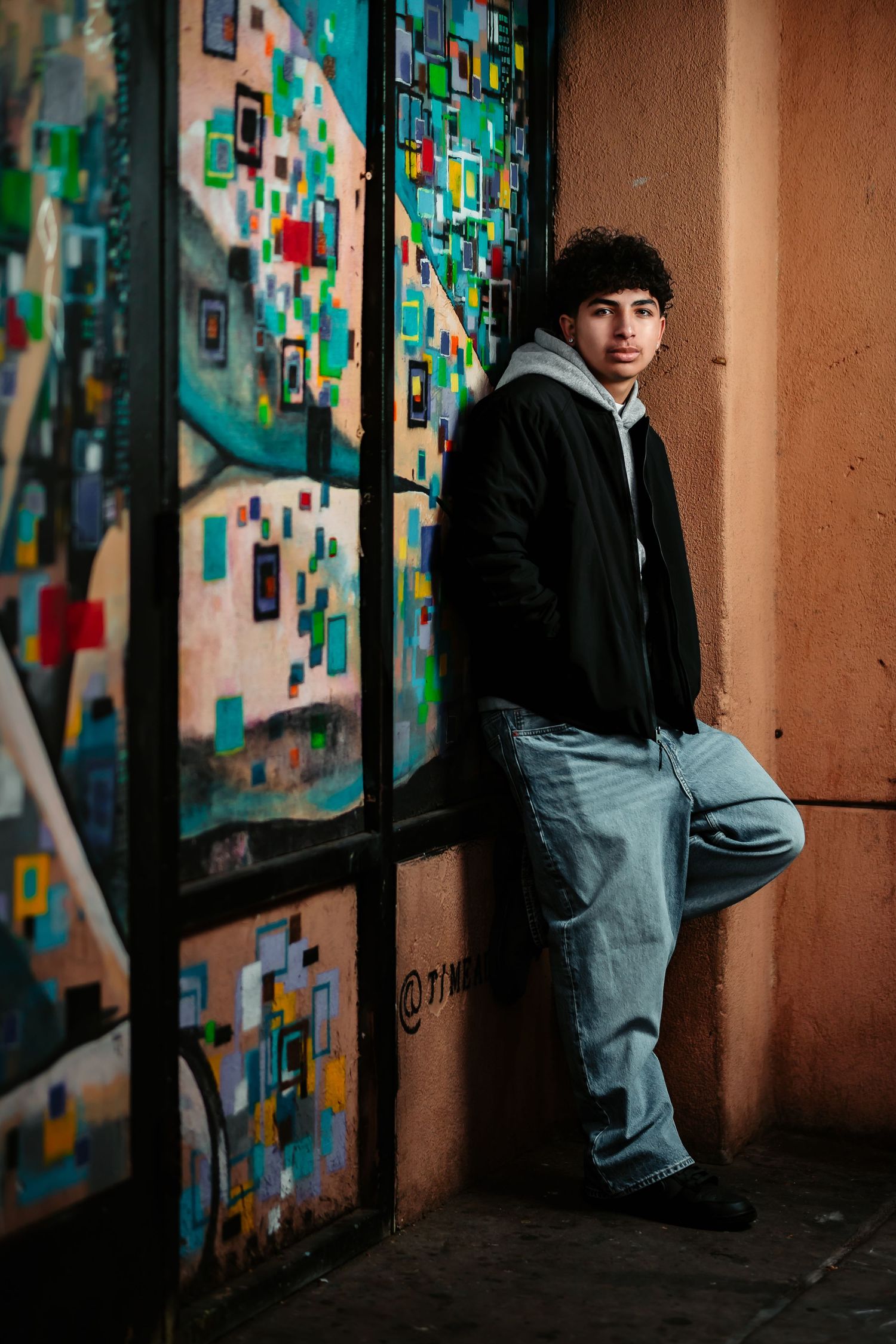 Person in black jacket and jeans sits against wall with colorful geometric mural art in dimly lit corridor.