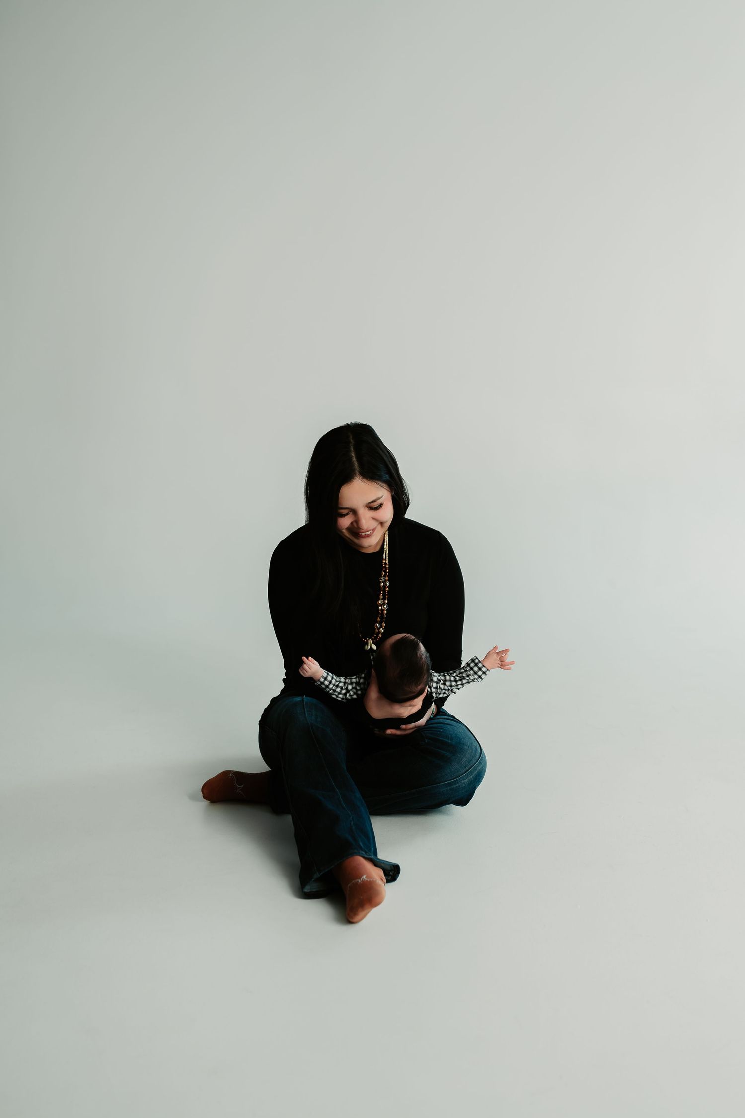 A person in black clothing sits curled up on a light gray floor in a minimalist setting.