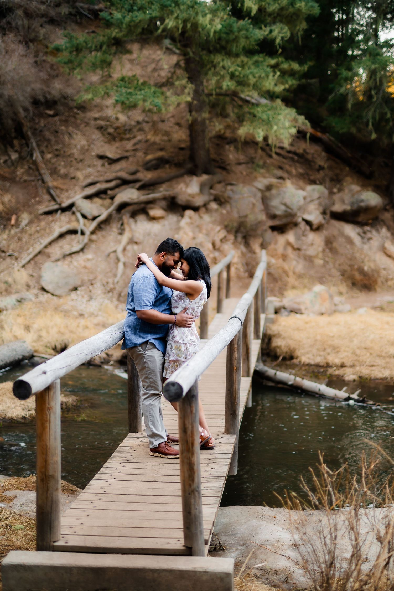 A couple embraces on a rustic wooden bridge with rocky cliffs and water in the background.