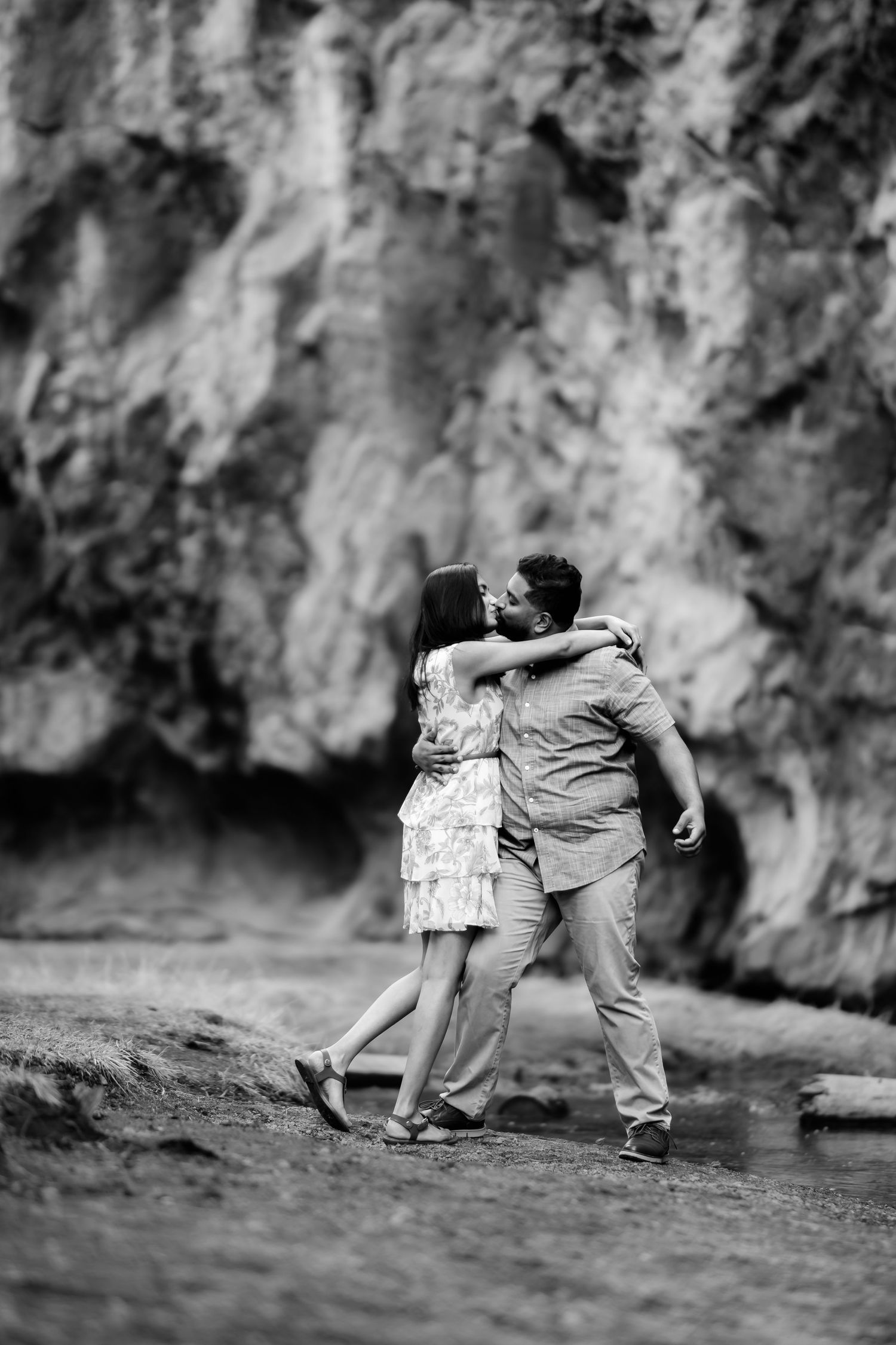 A romantic couple embraces while standing near rocky cliffs in a black and white nature scene.