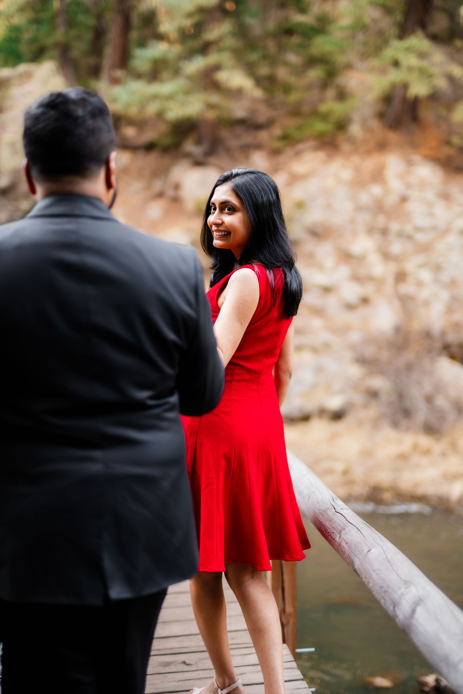 Couple stands on wooden deck overlooking mountain stream with rocky cliff backdrop.