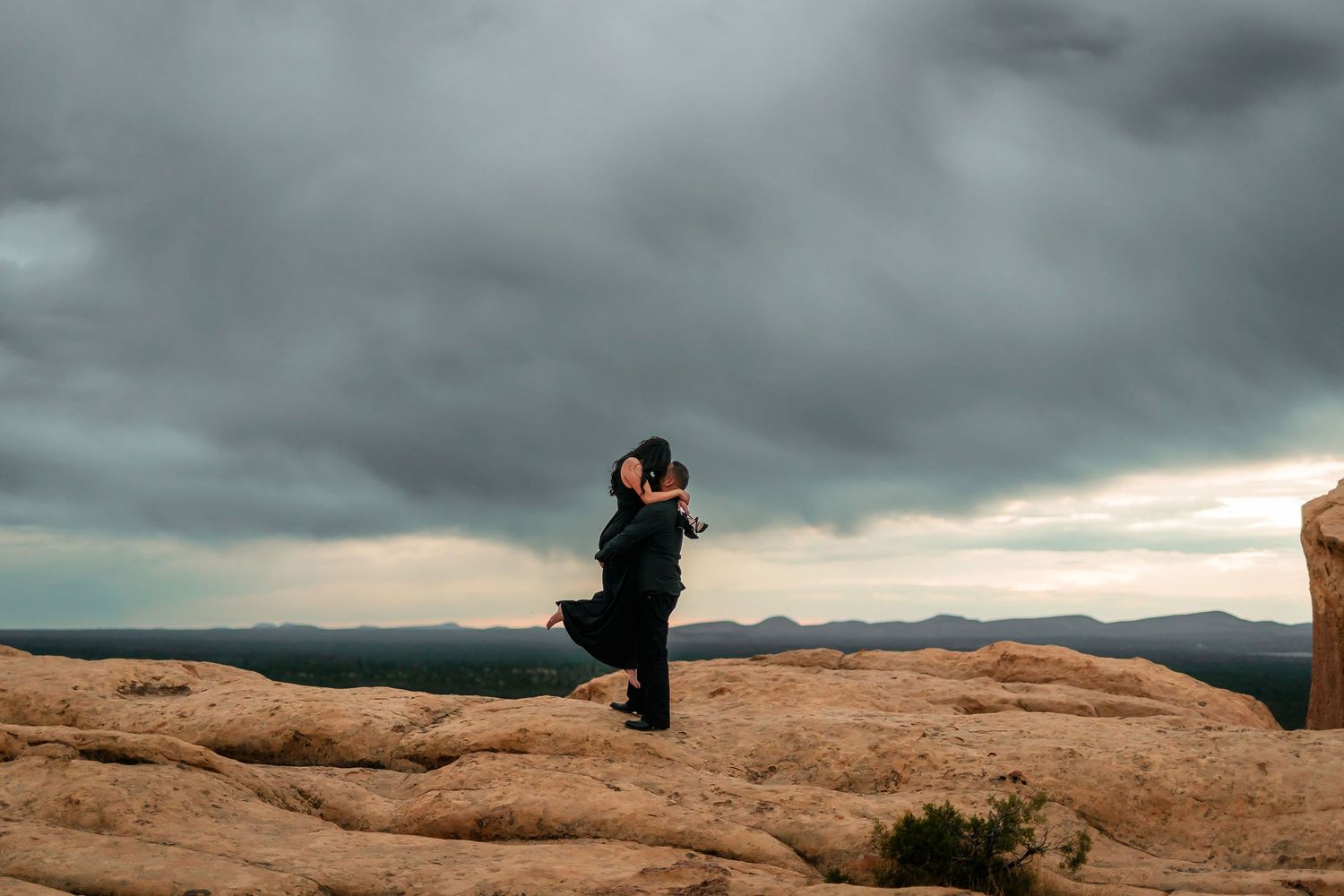 Couple embracing on rocky desert landscape under dramatic stormy sky with mountain views.