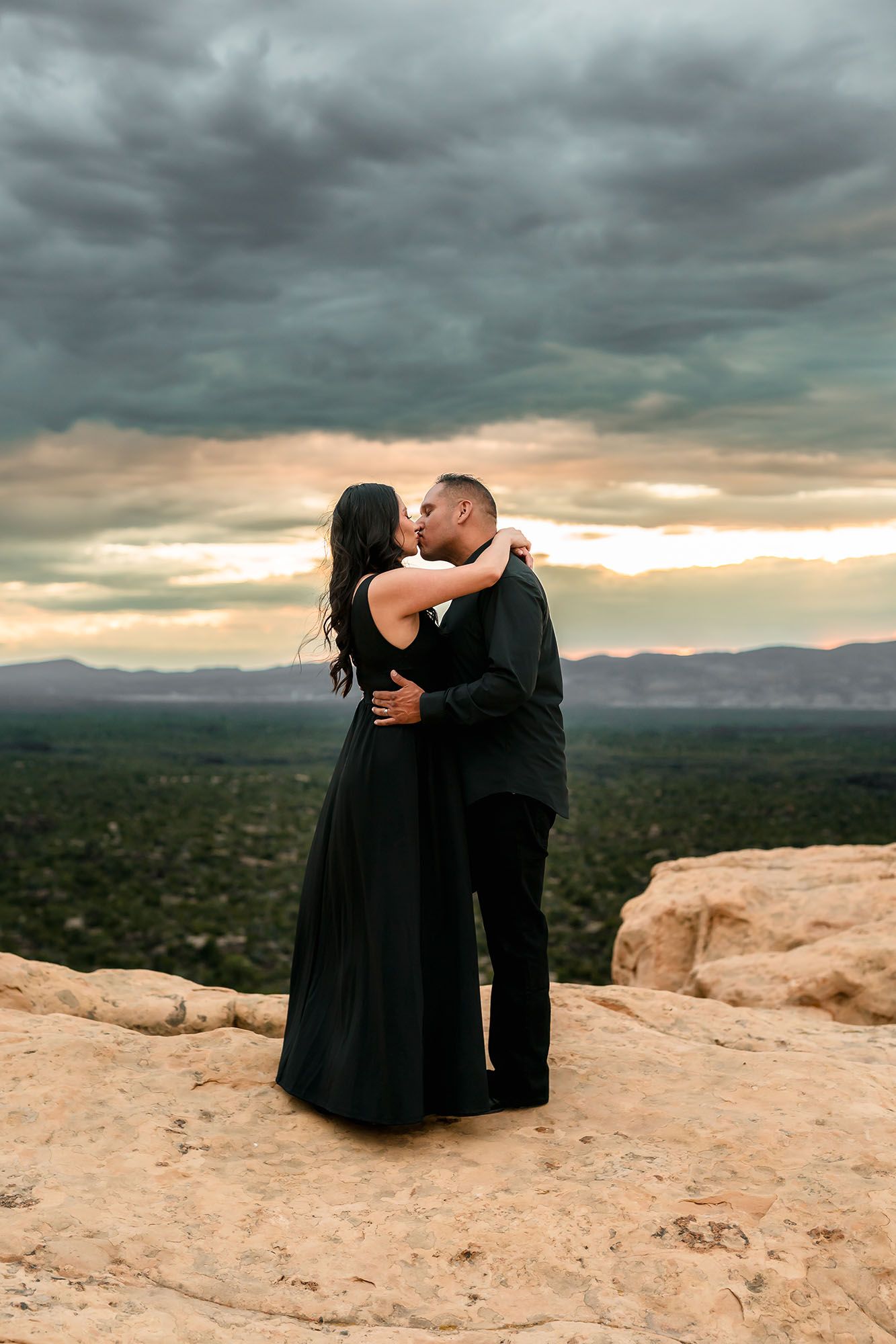 Couple kissing in matching black outfits on desert rock ledge with stormy sunset sky and valley below.