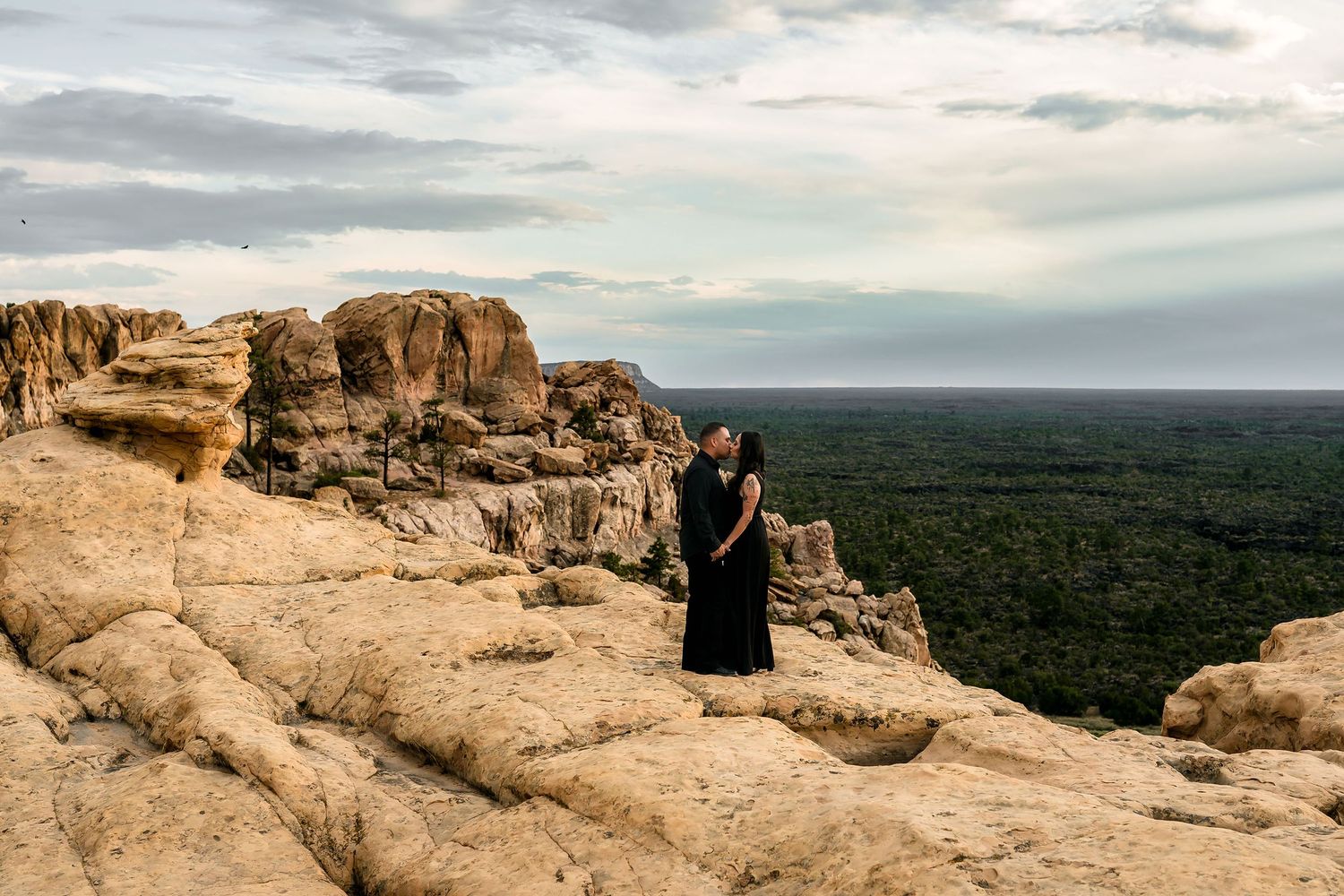 Couple in black embracing on rocky cliff edge with vast green landscape and cloudy sky backdrop.