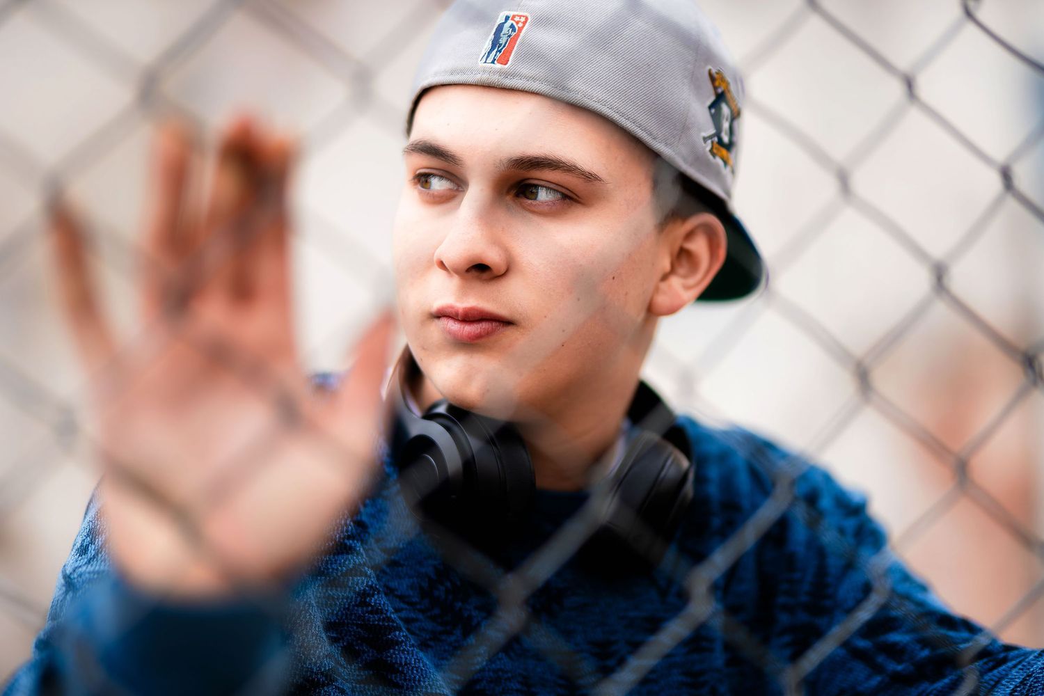 Young man in snapback cap and headphones pressing hand against chain-link fence outdoors.