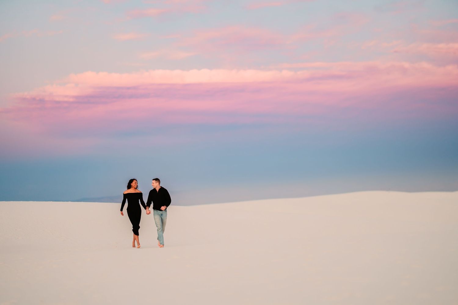 Two silhouettes walk together on a beach at sunset with pink and purple clouds in the sky.