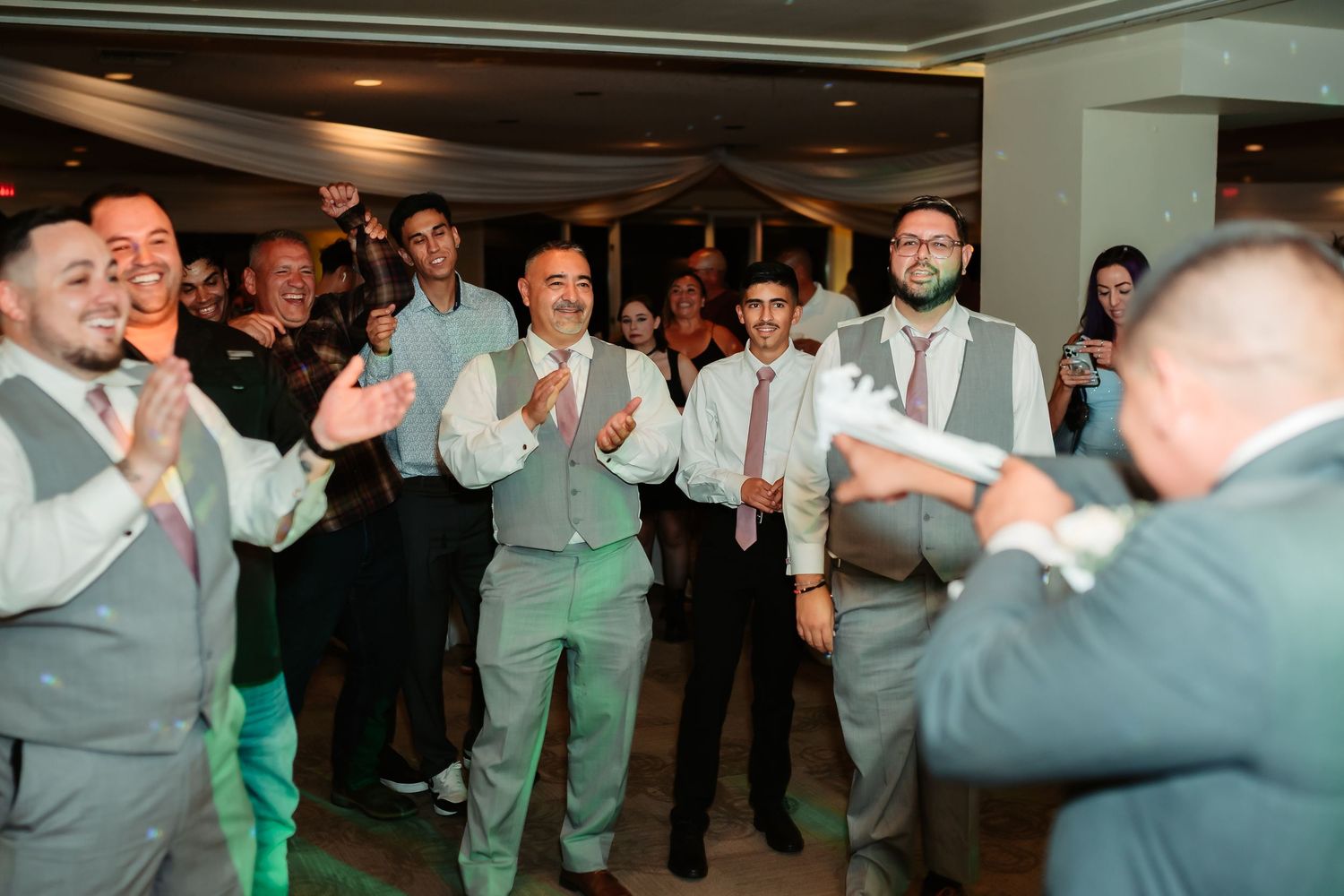 Groomsmen in matching grey vests and ties celebrating enthusiastically at an indoor wedding reception.