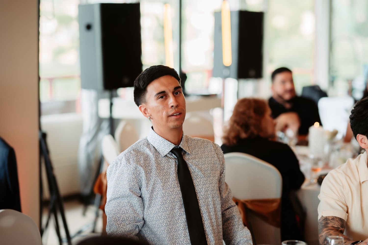 A guest in a white dress shirt and tie sits attentively at an indoor event with speakers in the background.