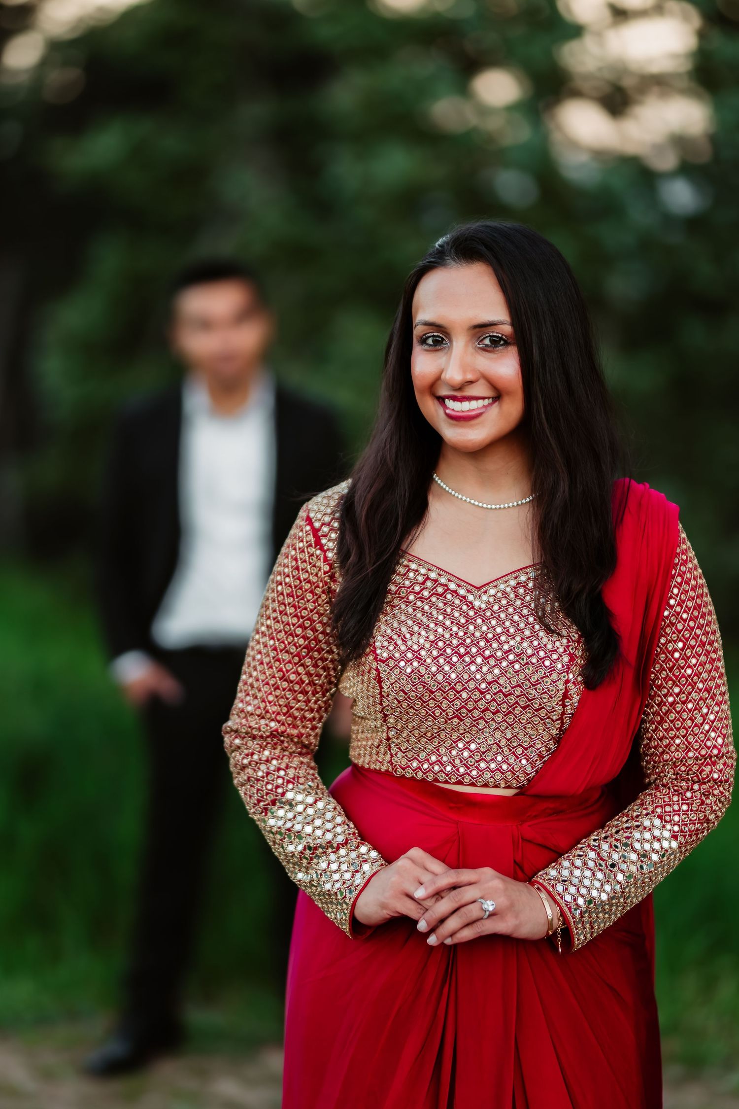 Person in traditional Indian formal attire poses outdoors in red and gold embellished dress against natural background.