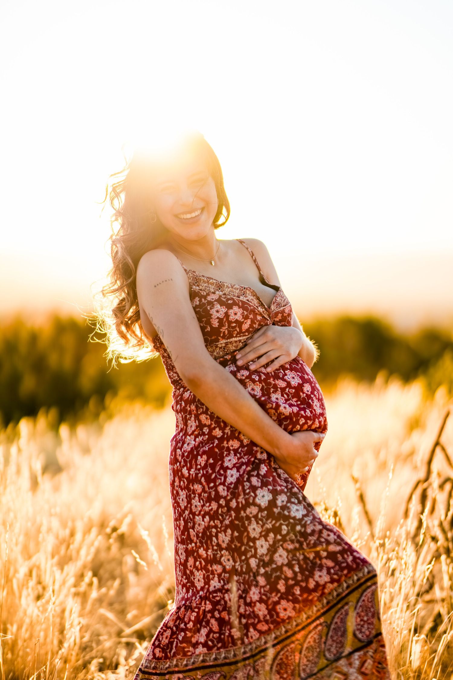Pregnant woman in floral dress cradles her belly while standing in golden wheat field at sunset.